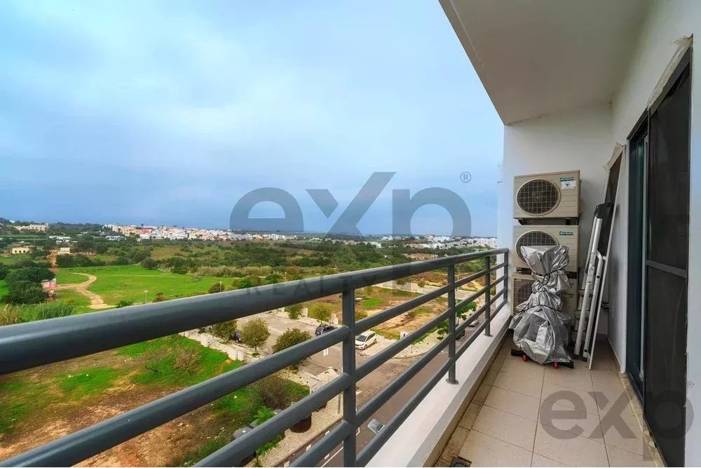 Balcony with metal railing overlooking green fields and distant buildings under a cloudy sky; air conditioning units and covered equipment on the wall to the right.
