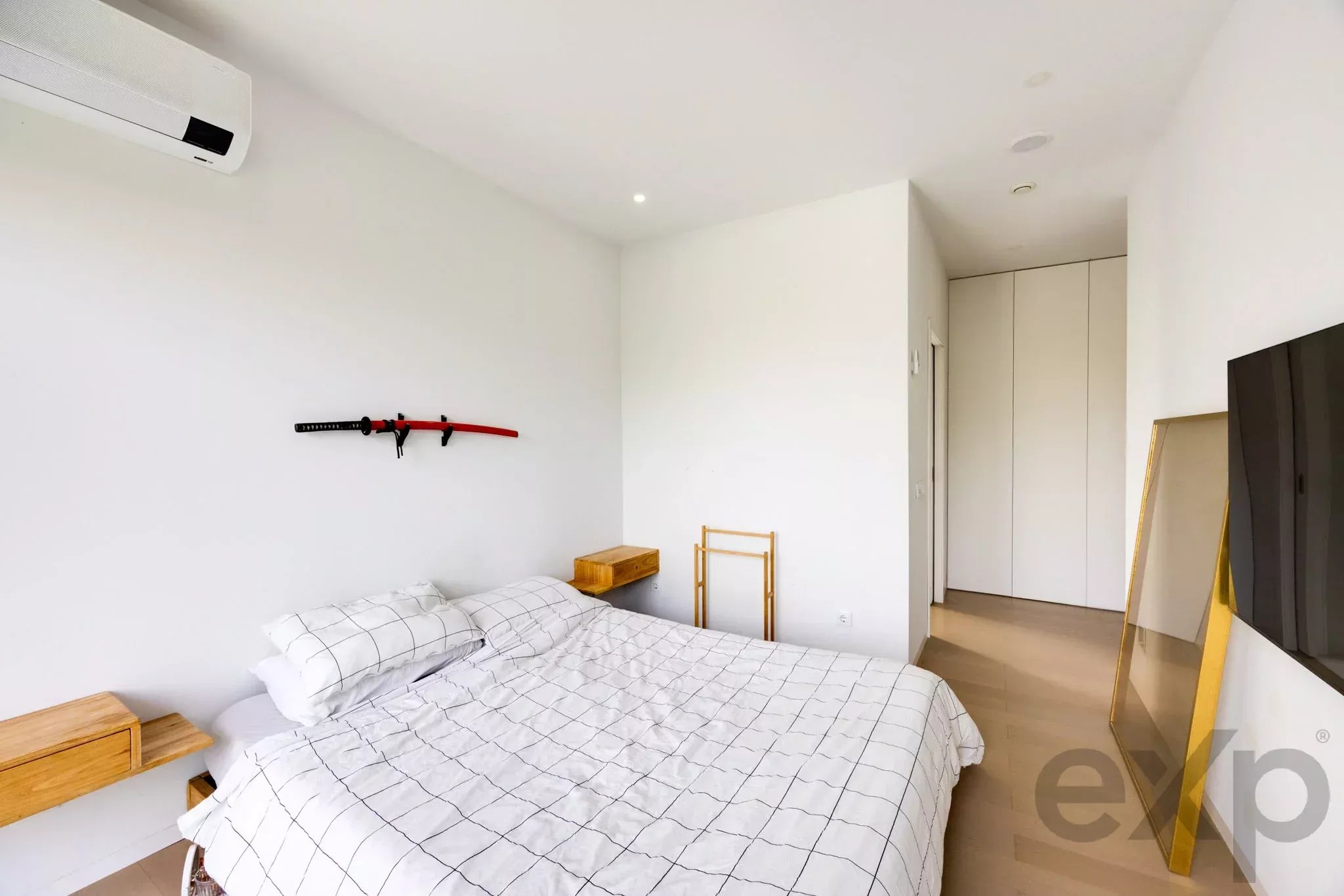Minimalist bedroom with white walls, a bed with grid-pattern bedding, a wall-mounted red-and-black katana, a small wooden nightstand, and built-in cabinets.