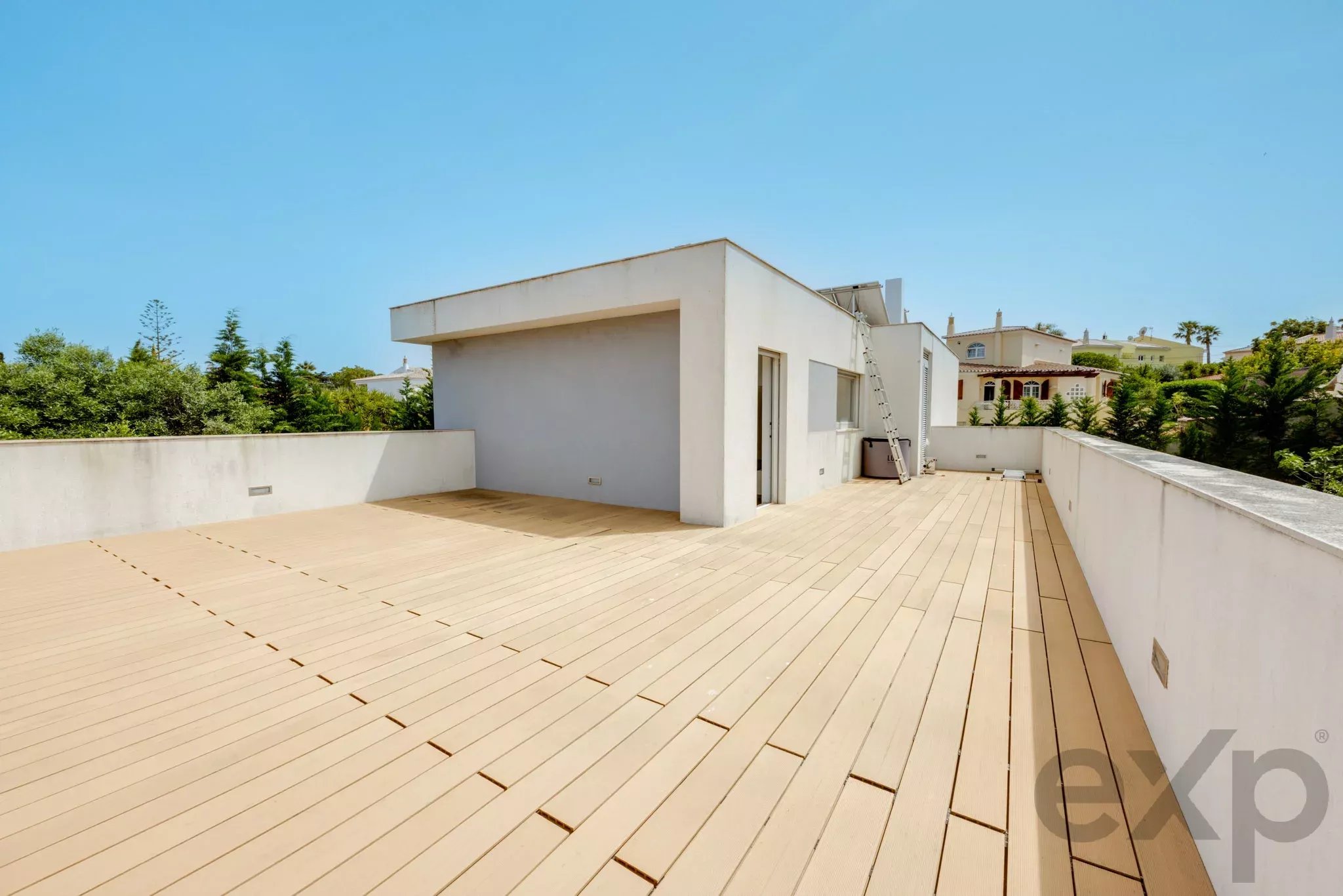 Rooftop terrace with light wooden decking, white walls, and a bright blue sky above.
