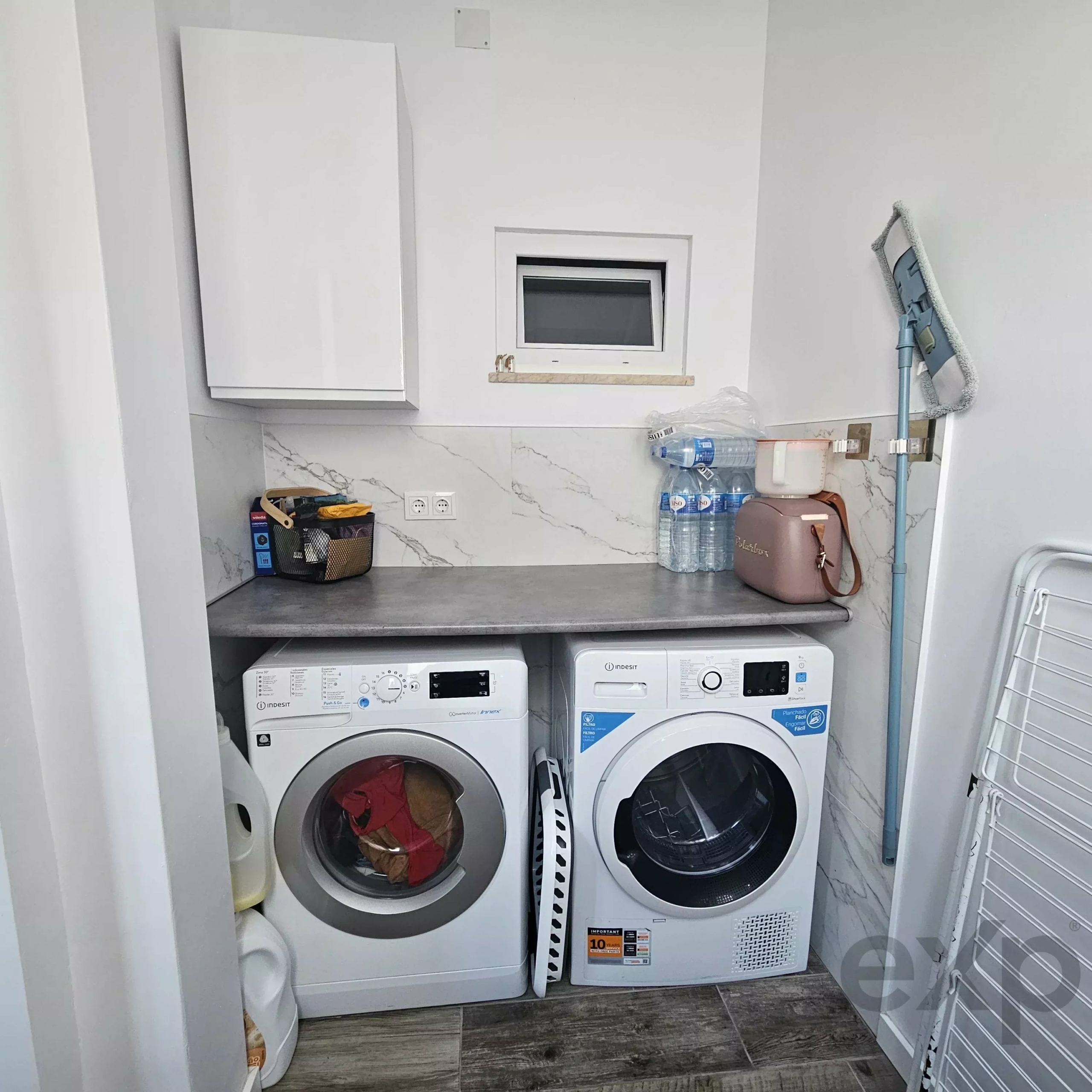 Laundry closet with two front-loading appliances (washer and dryer) and white cabinets above, clutter including detergent and water bottles.