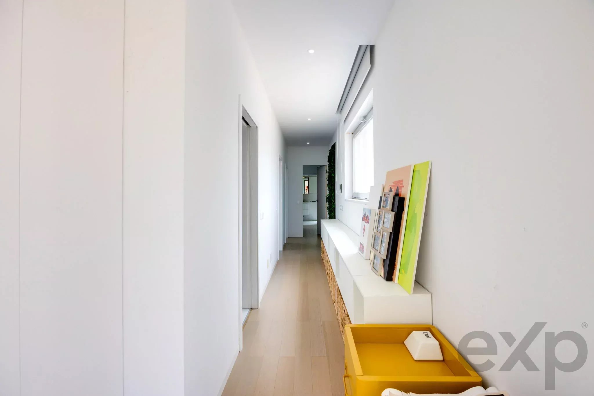 Bright white hallway with a white console on the right displaying colorful art prints; a yellow crate sits on the floor in the foreground.
