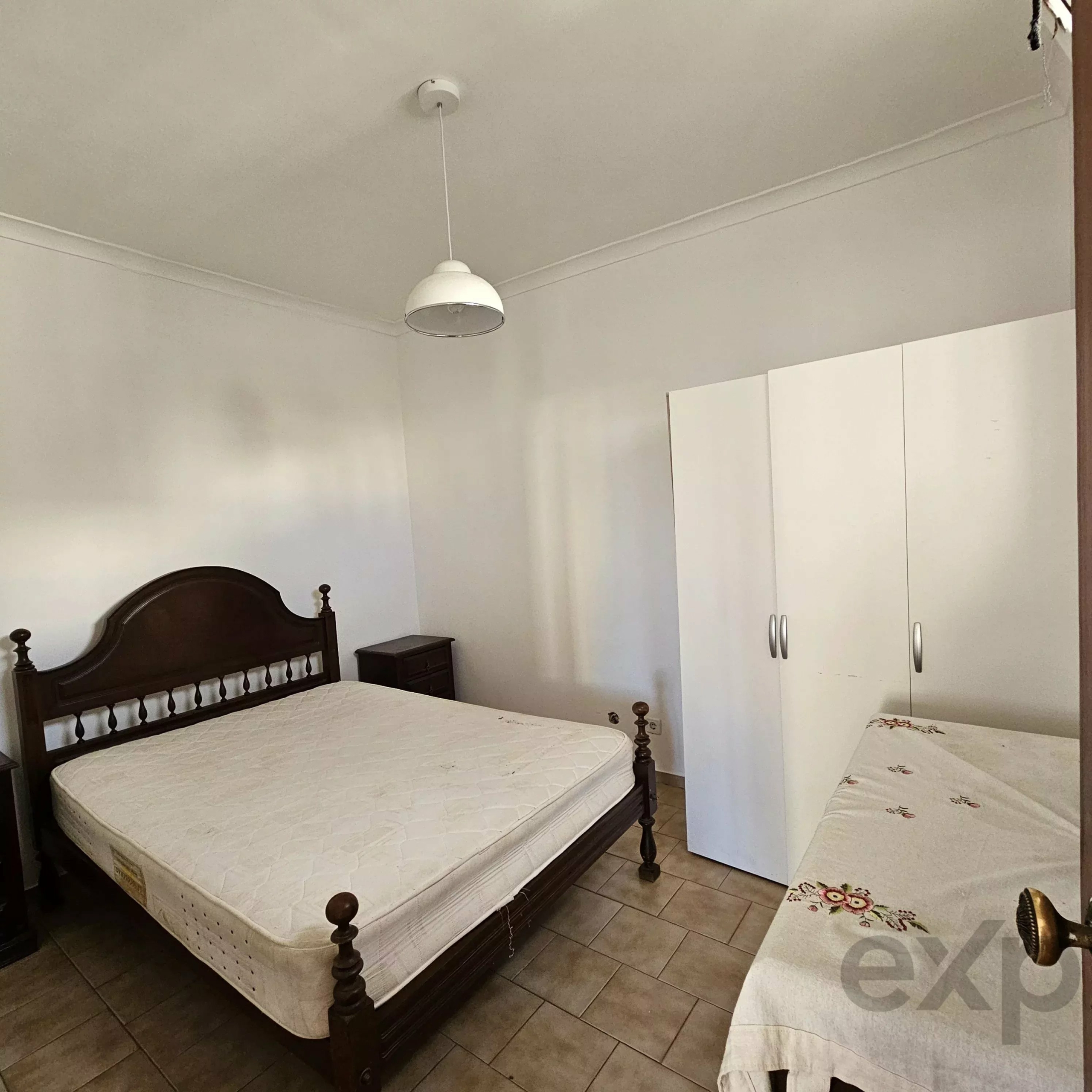 Bedroom with a dark wooden bed frame and two mattresses, a white pendant ceiling light, and white storage cabinets along the wall; tiled floor is visible.