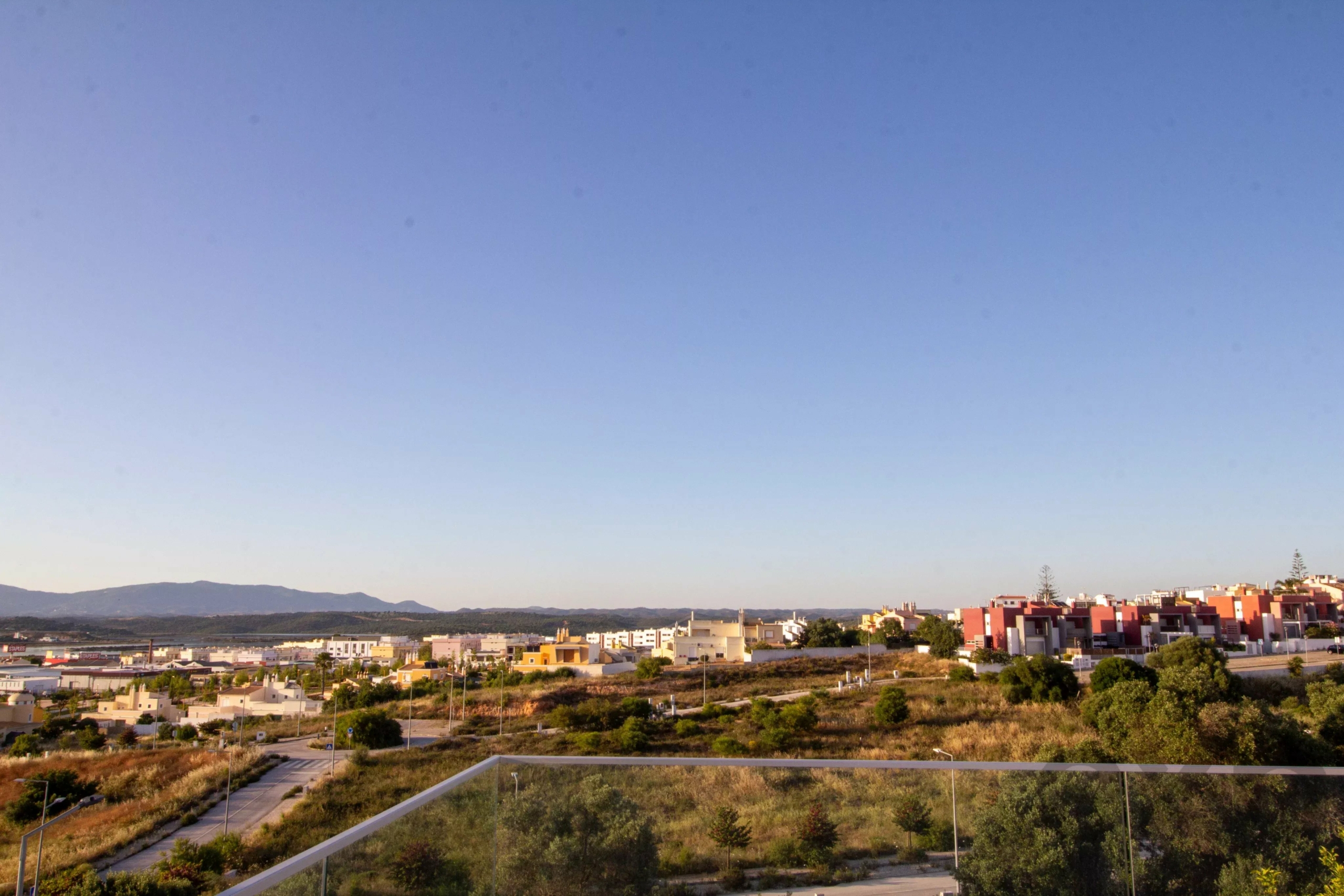 Balcony glass railing in the foreground overlooking a suburban hillside neighborhood with low-rise buildings and distant hills under a clear blue sky.
