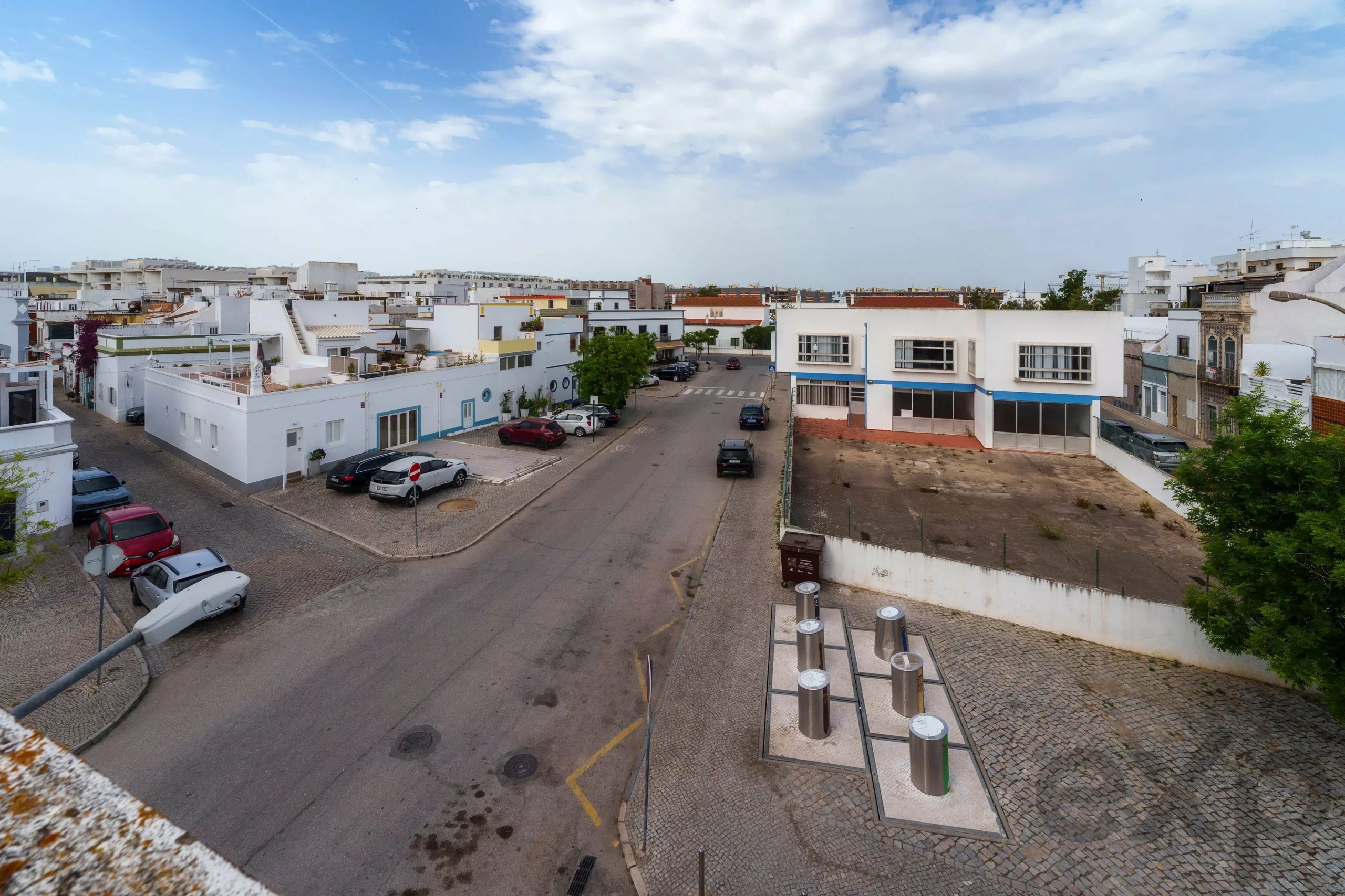 Aerial view of a sunny street in a white-washed coastal town with parked cars and low-rise buildings.