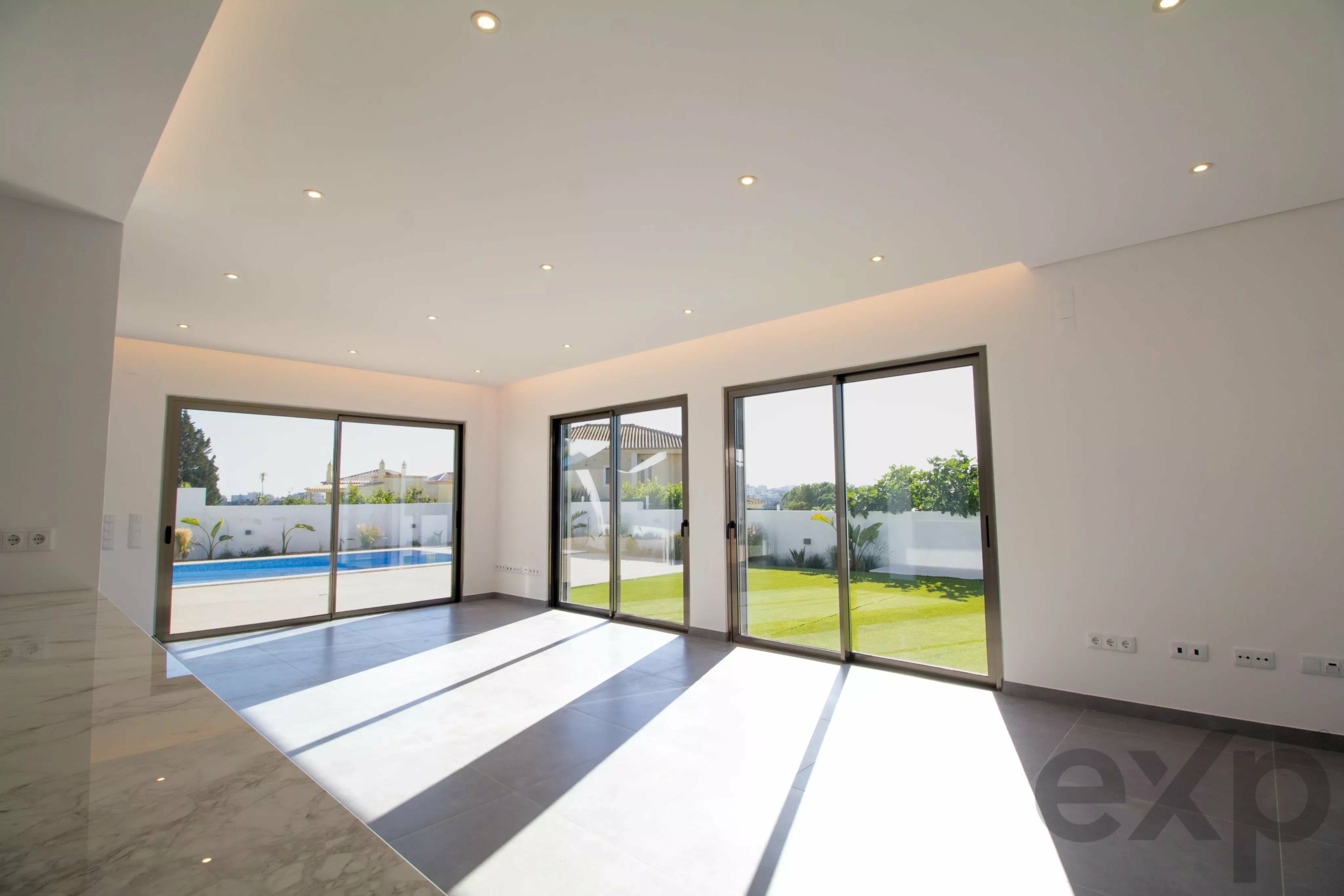 Bright modern living area with huge glass sliding doors overlooking a pool and garden outside; white walls, marble floor, and recessed ceiling lights.