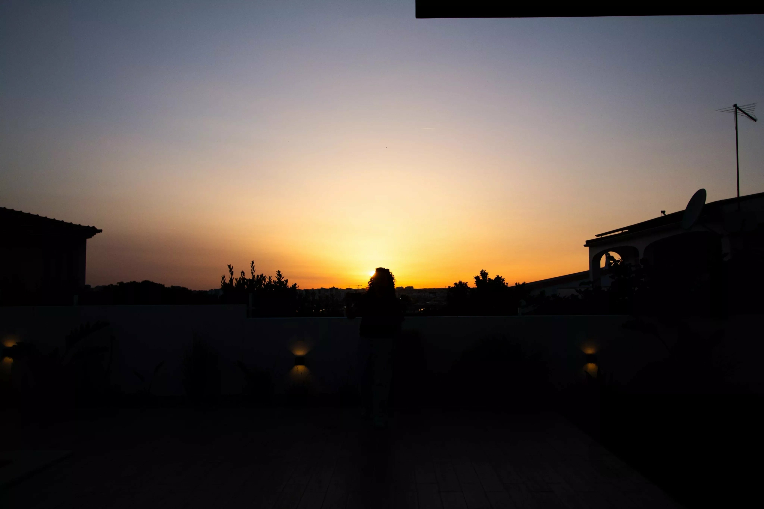 Silhouette of a person taking a photo on a rooftop at sunset, with antennaes and dark buildings along the horizon.