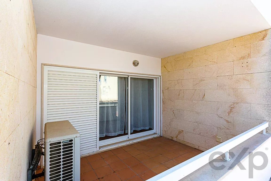 Narrow balcony with terracotta floor tiles, white railing in the foreground, sliding glass door with a closed shutter, and an outdoor AC unit on the left.