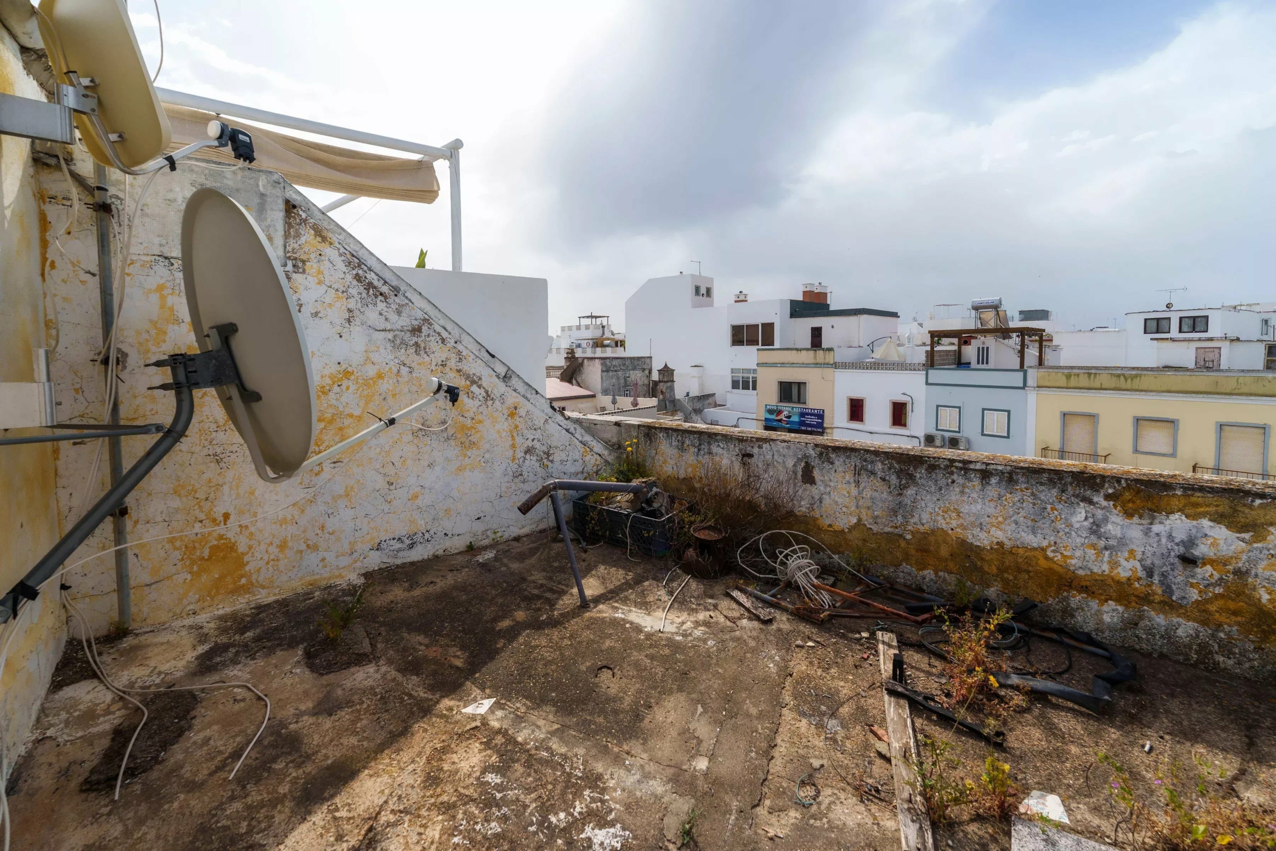 Rooftop with a satellite dish and cables on a weathered wall, overlooking white buildings under a cloudy sky.
