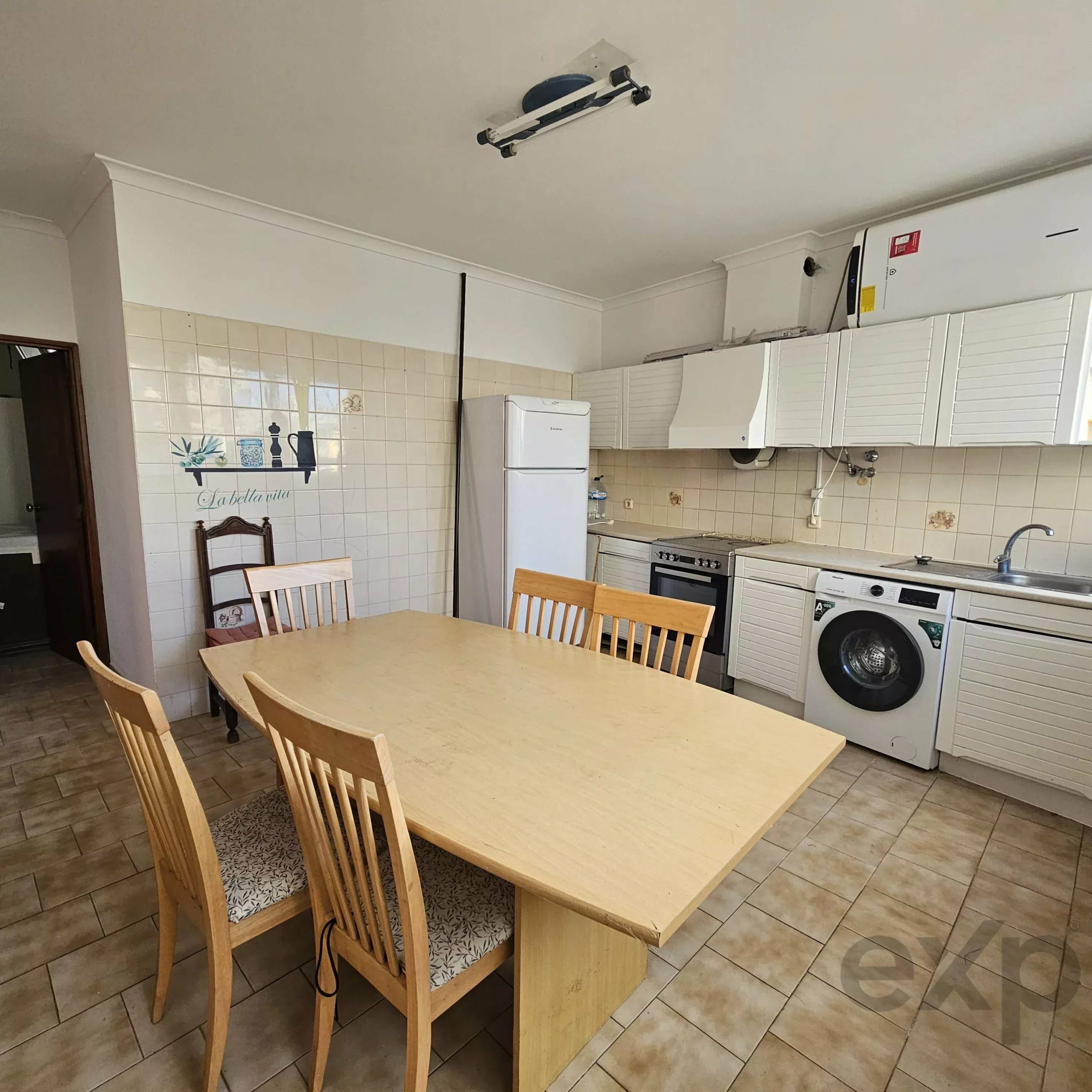 A light wooden dining table with matching chairs in a tiled kitchen area, white cabinets and appliances visible in the background.