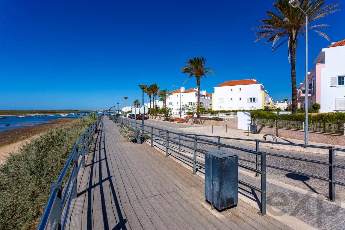 Boardwalk along the coast with white houses, palm trees, and a bright blue sky on a sunny day.