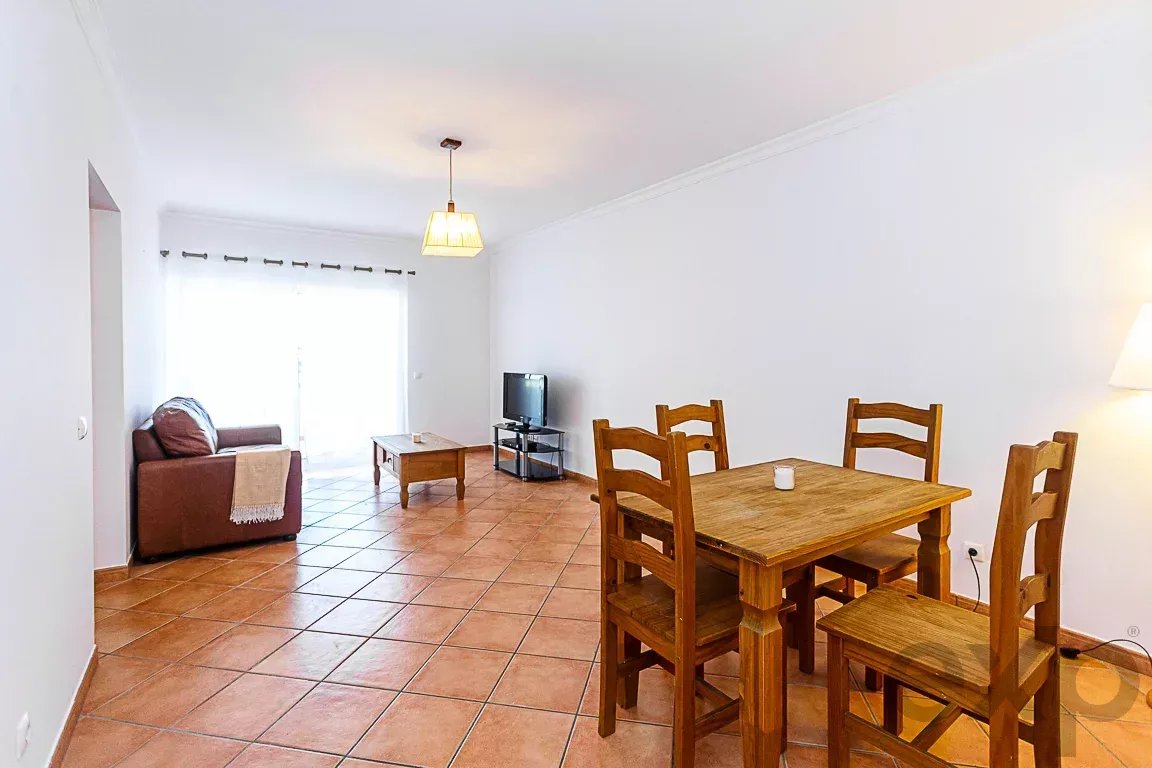 Open living and dining area with a wooden dining table and four chairs, a brown sofa, coffee table, and a small TV on a stand.