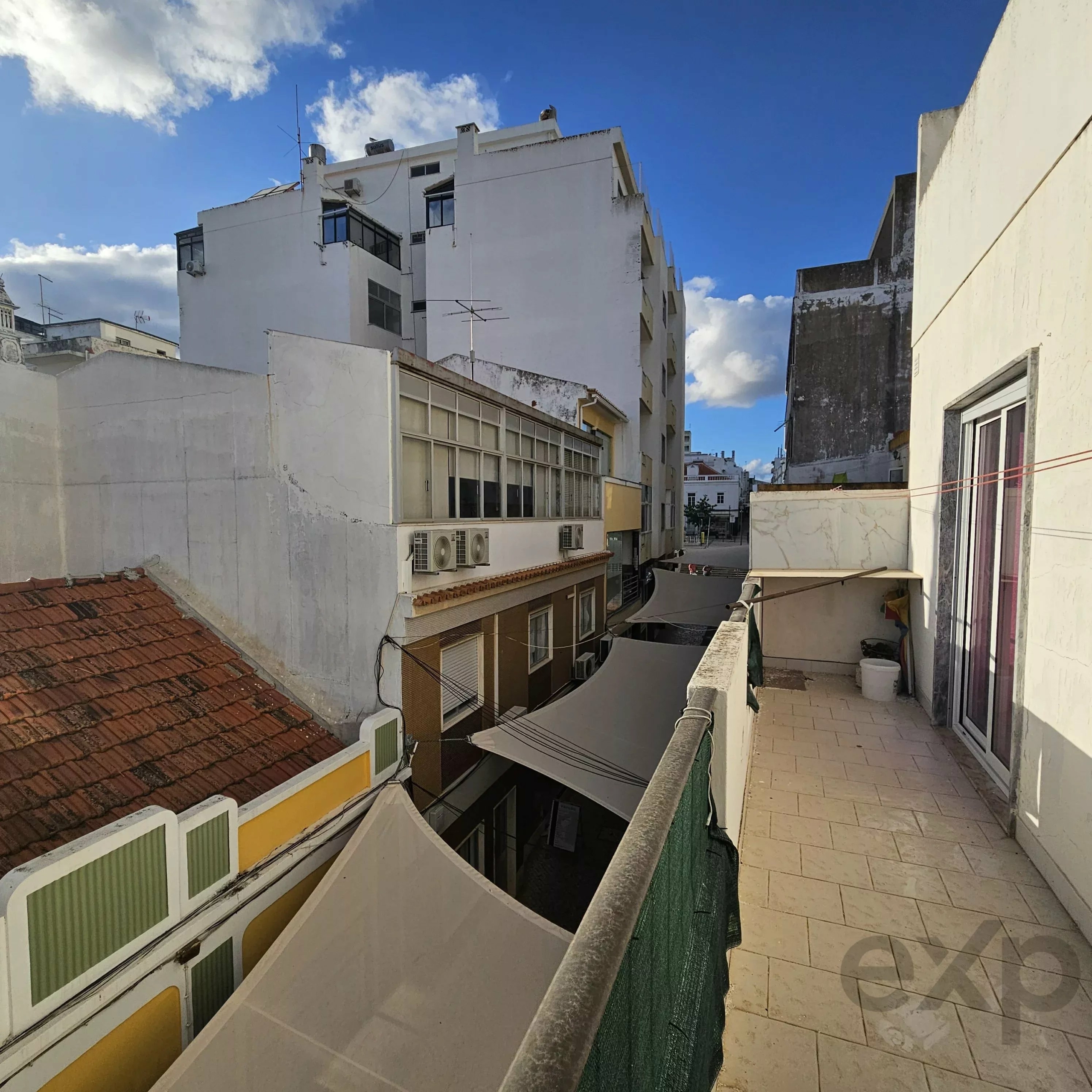 Narrow alley between white apartment buildings under a bright blue sky, with railing in the foreground and balconies above.