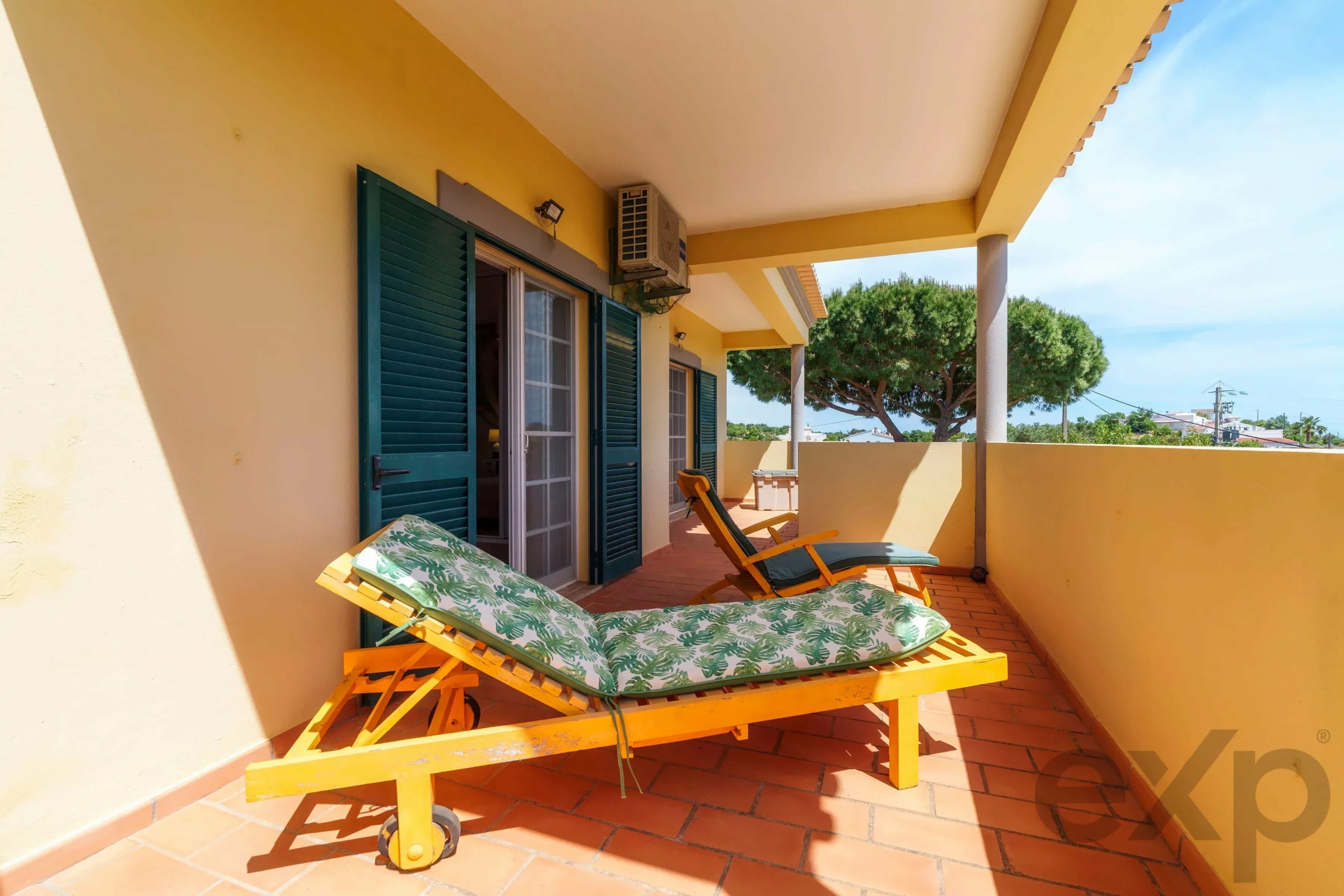 Sunny balcony with yellow walls, green shutters, and two wooden lounge chairs with palm-pattern cushions on terracotta tiles.