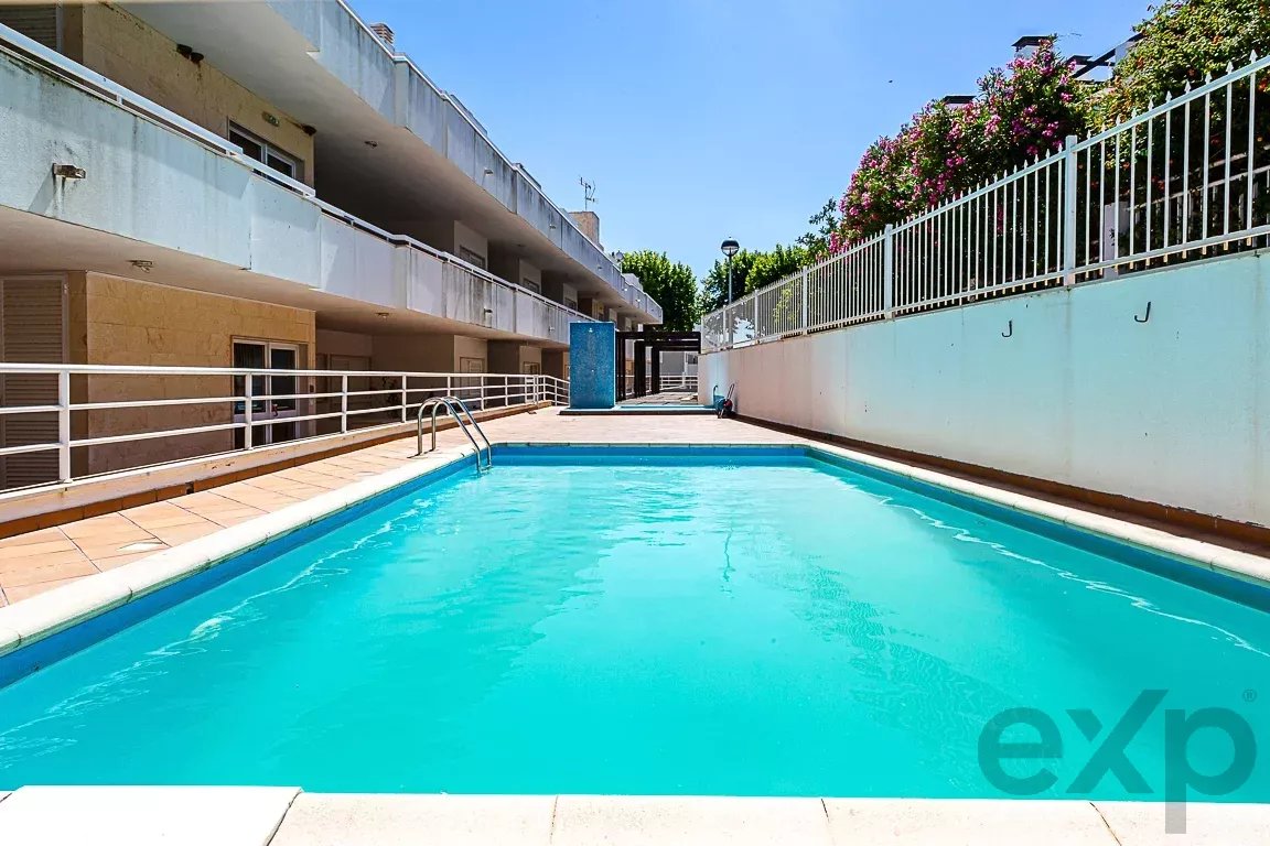Rectangular outdoor swimming pool with turquoise water, beige tile deck, and white railings beside a low-rise apartment building under a blue sky.