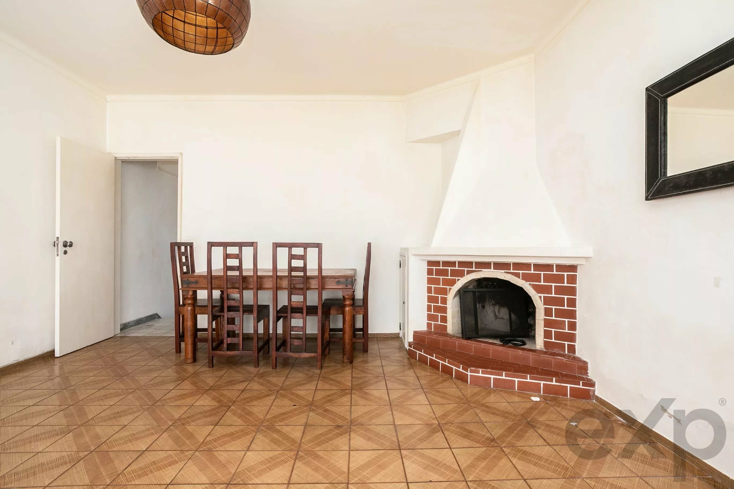 Dining area with a wooden table and six chairs, a brick fireplace, and a doorway on the left in a bright, minimalist room.