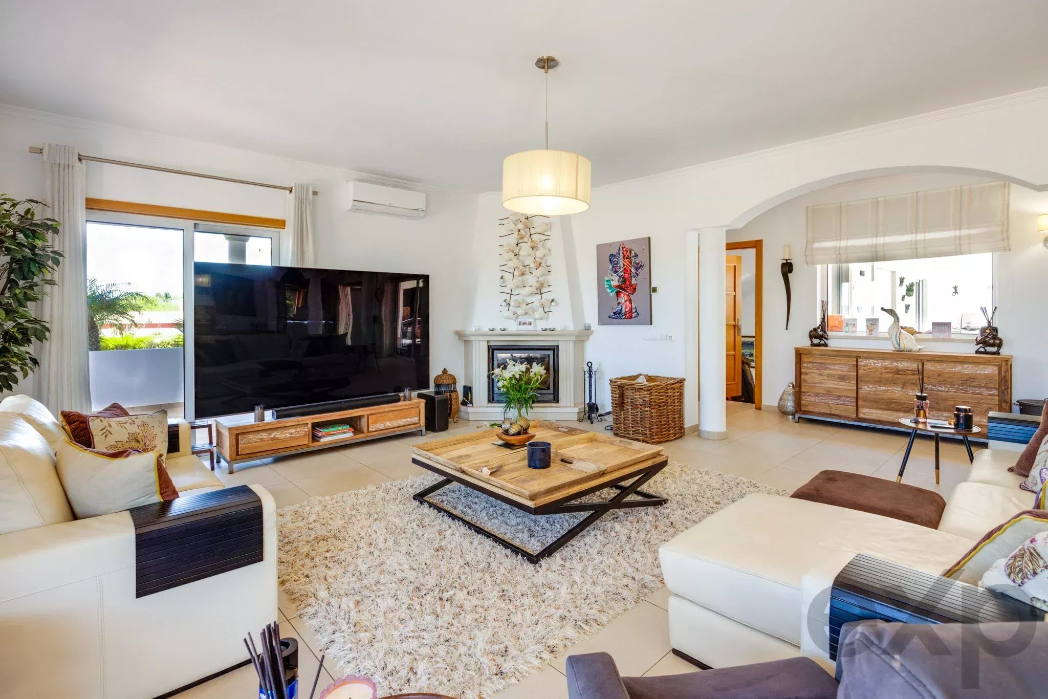 Bright living room with white sofas, a large flat-screen TV, and a stone fireplace under a hanging ceiling light.