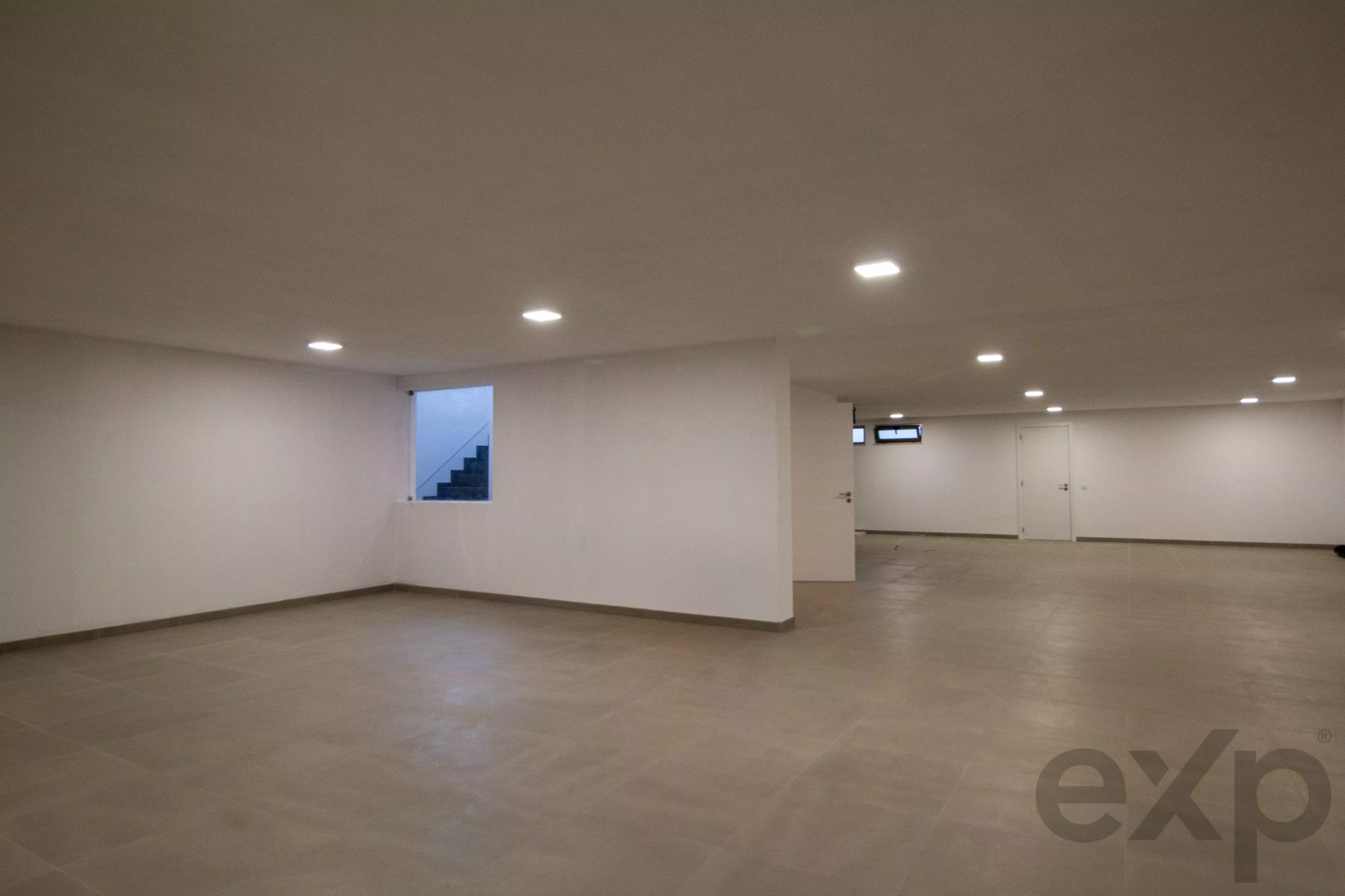 A large empty indoor room with white walls, grey tiled floor, and evenly spaced ceiling lights; a stairwell is visible through a small window in the wall.
