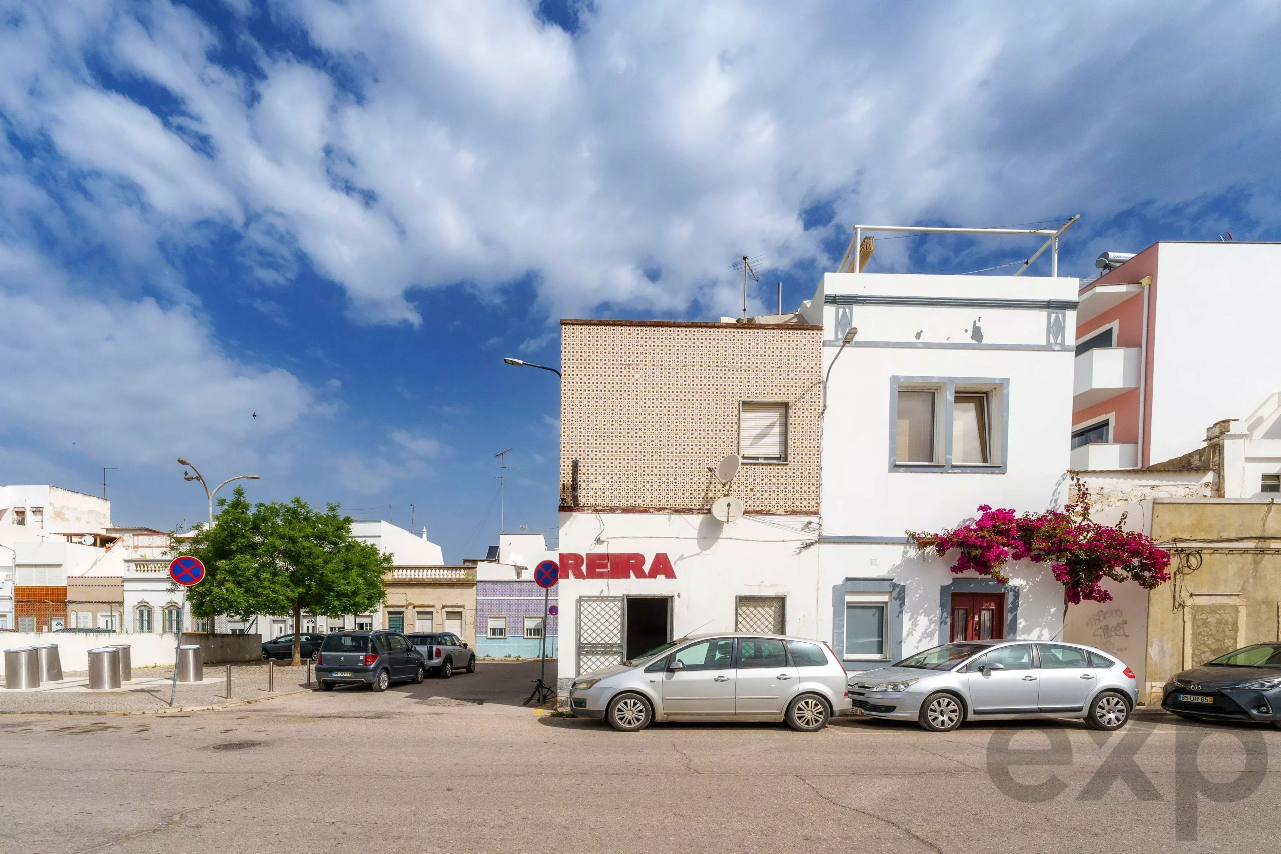 Urban street scene with white buildings, parked cars, and a partly tiled wall against a blue, cloudy sky.