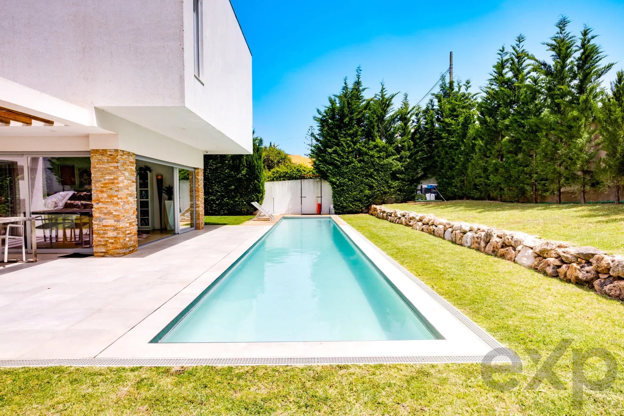 Modern white house with glass doors, stone column, and a long rectangular pool along a patio bordered by green lawn and tall evergreen trees under a clear blue sky.
