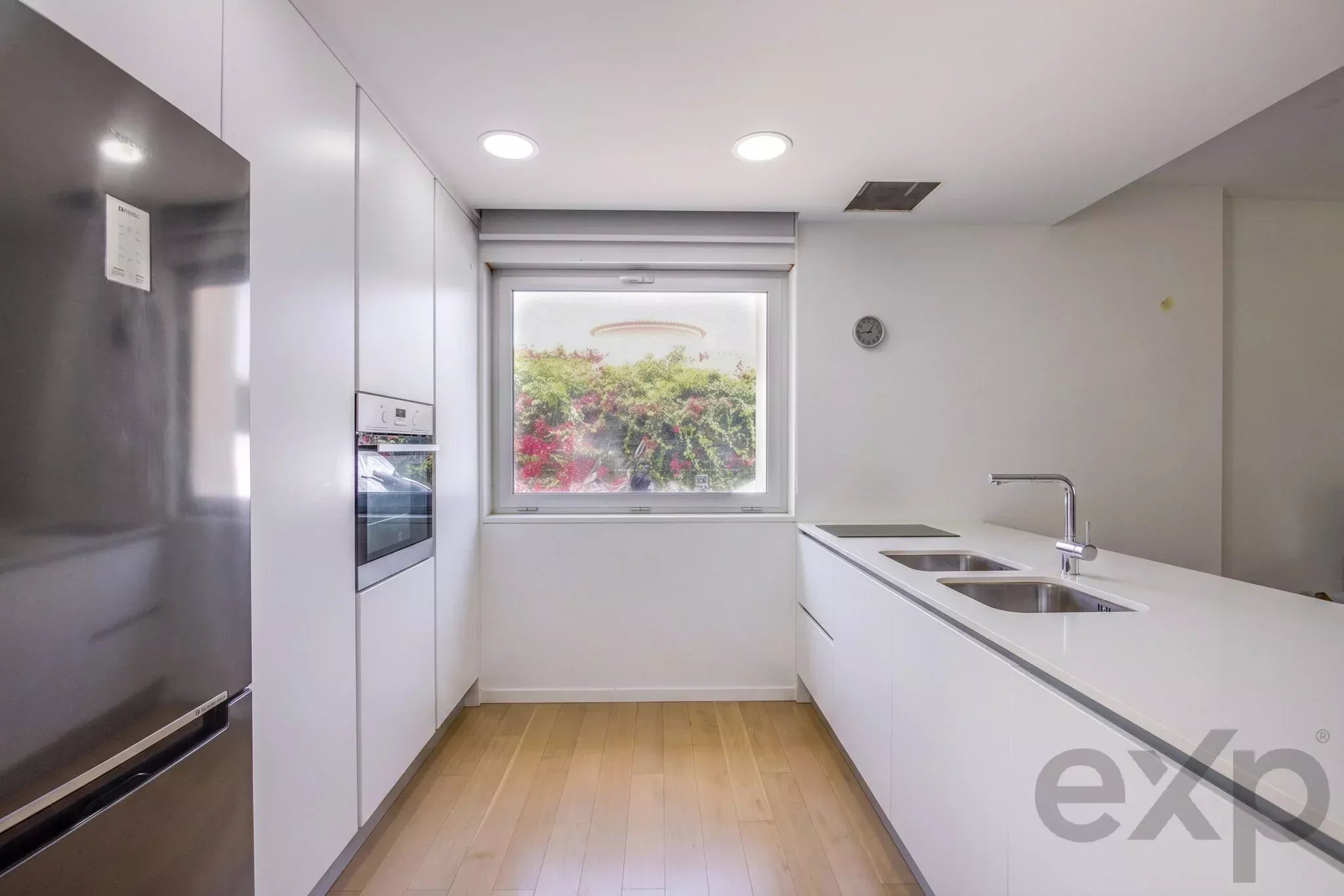 Bright white galley kitchen with a stainless steel fridge on the left, built-in oven, and a double sink on a white island.