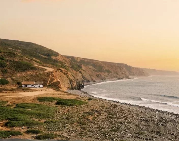 Sunset over a rocky coastal cliff with a dirt road and a parked bus along the shore.
