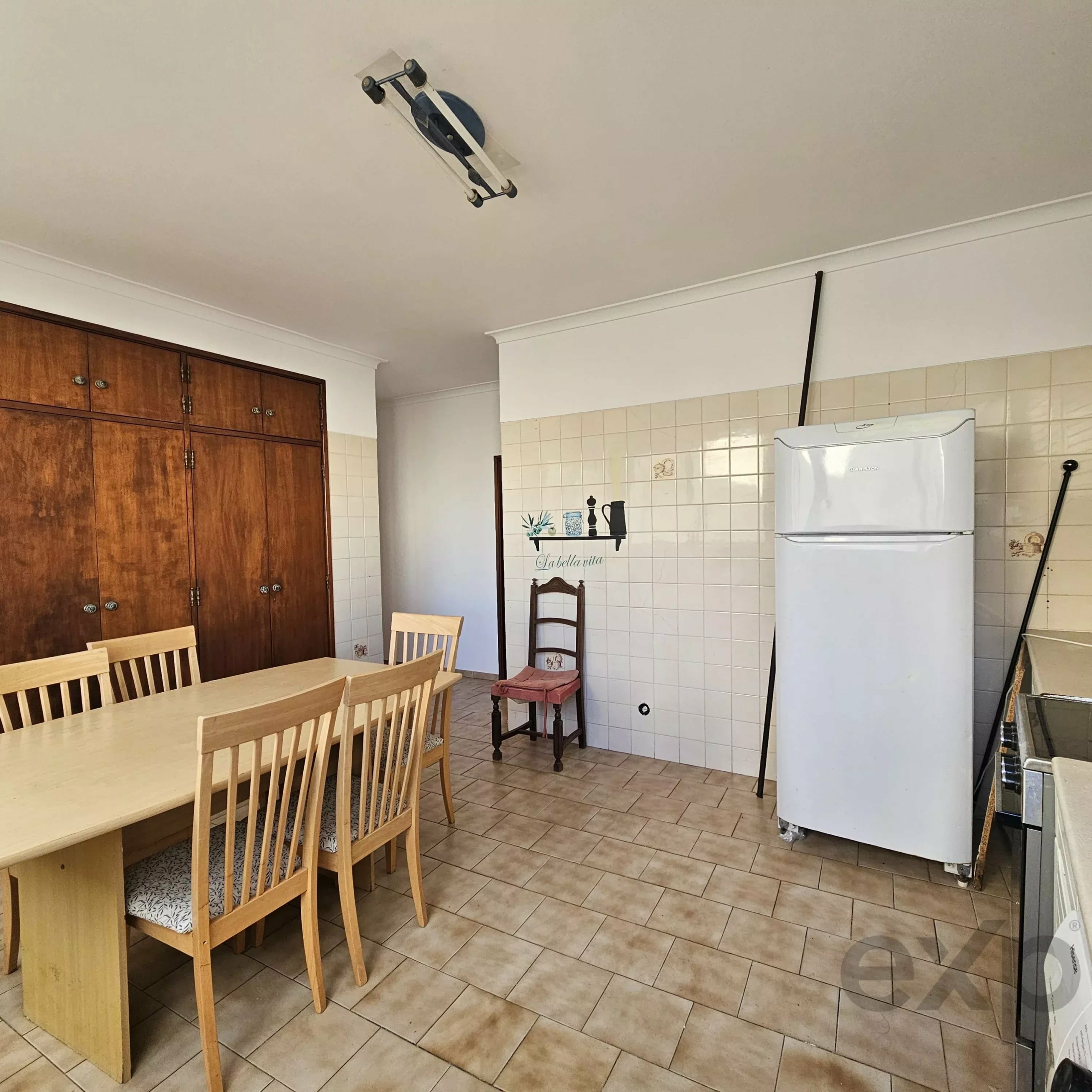 Wooden dining table and chairs in a kitchen, with tall wooden cabinets on the left and a white fridge on the right.