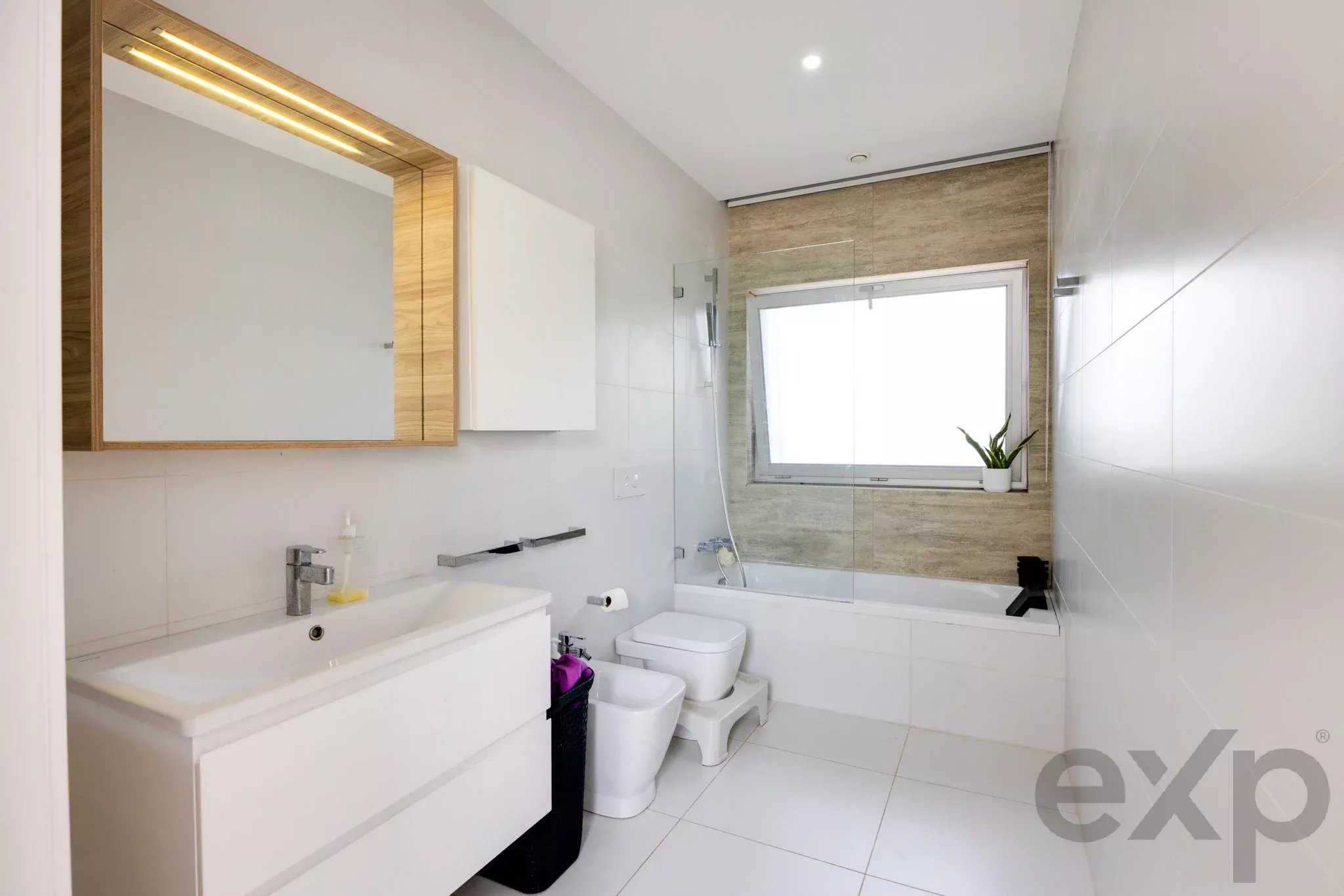 Modern bathroom with white vanity, large mirror, wall cabinet, toilet, bidet, glass shower, and a window with a potted plant on the sill.