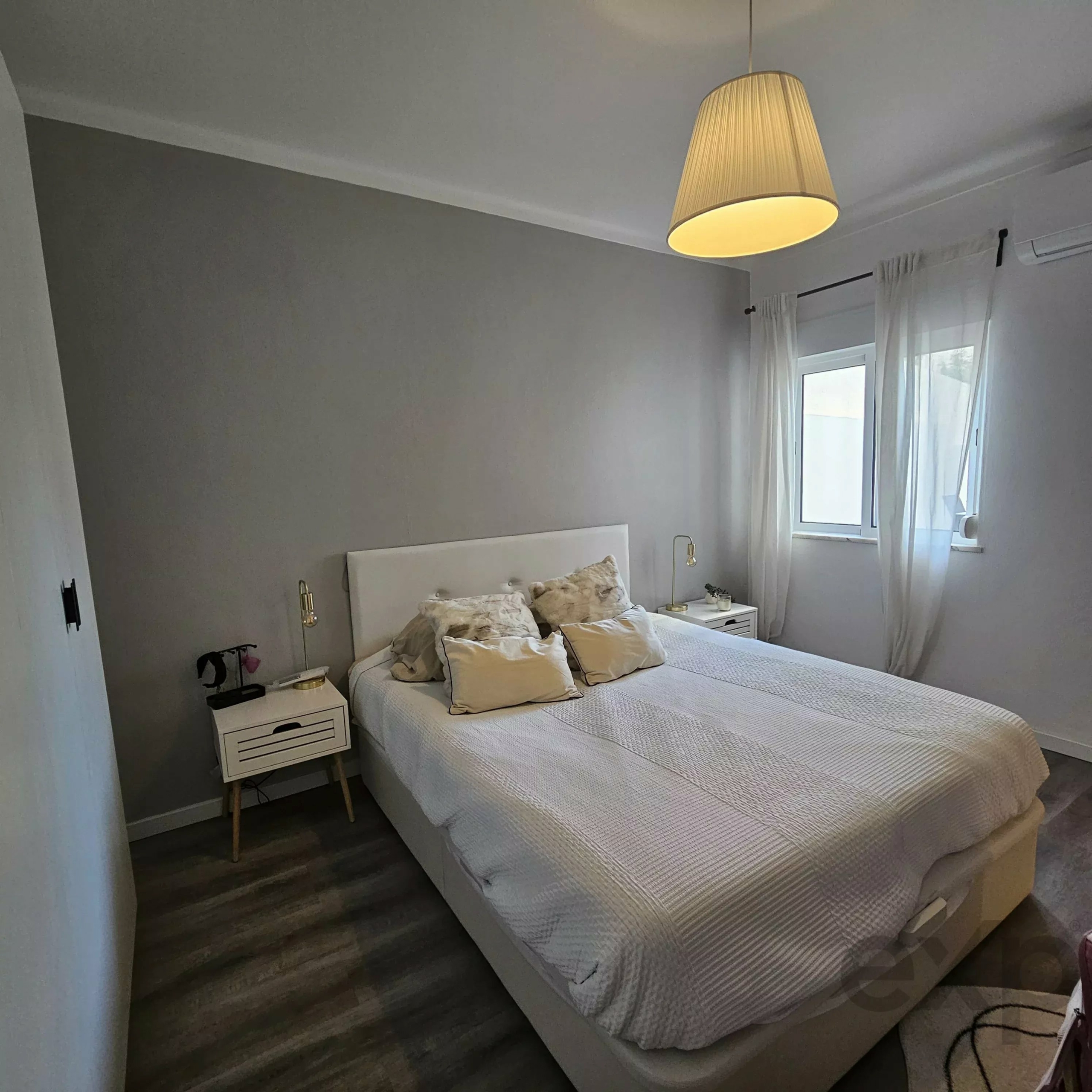 Bedroom with two single beds, white bedding, matching nightstands, and a hanging lamp; gray walls and sheer curtains by a window.
