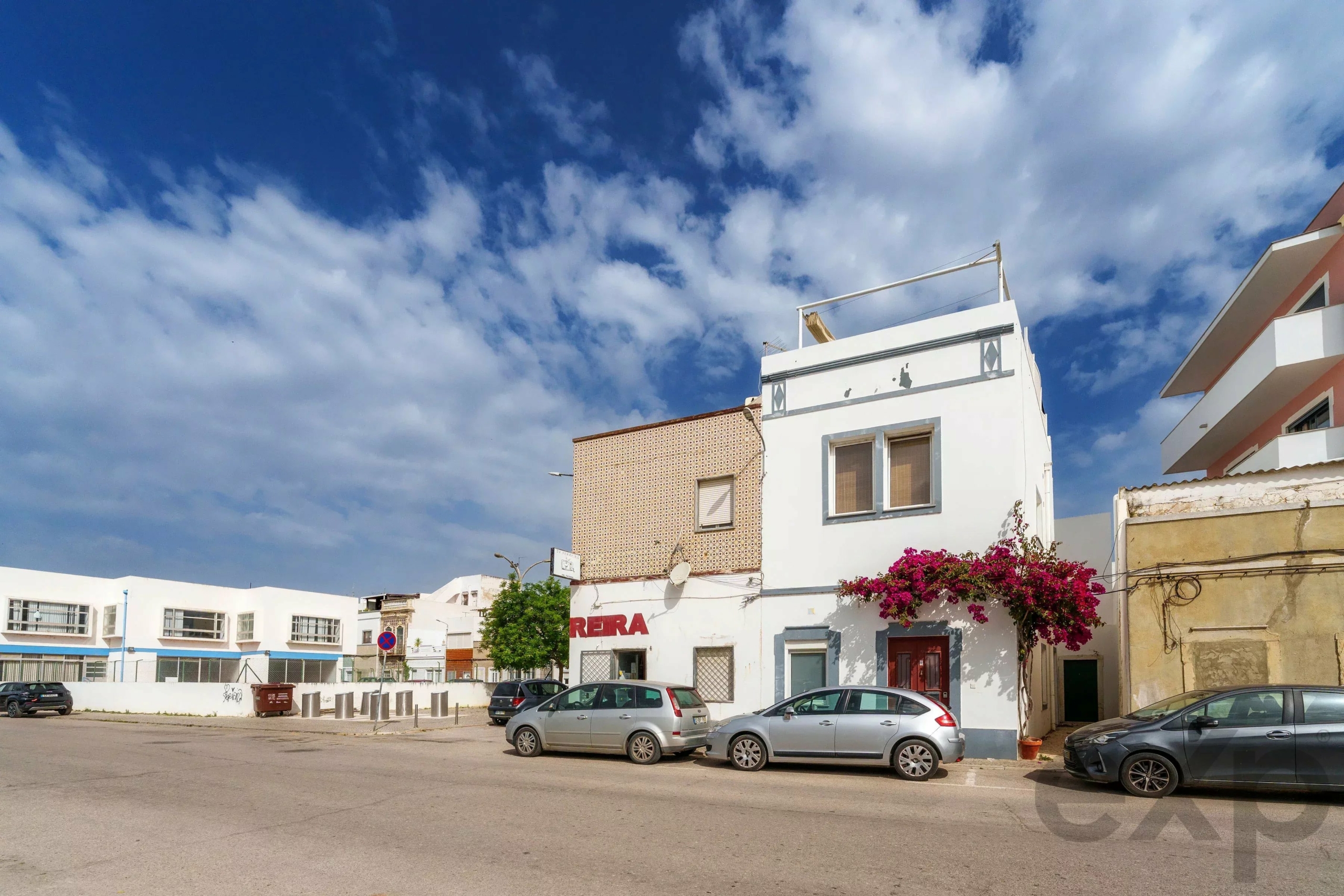 Urban street scene with parked cars in front of white buildings under a blue sky with clouds; pink bougainvillea growing beside the building.