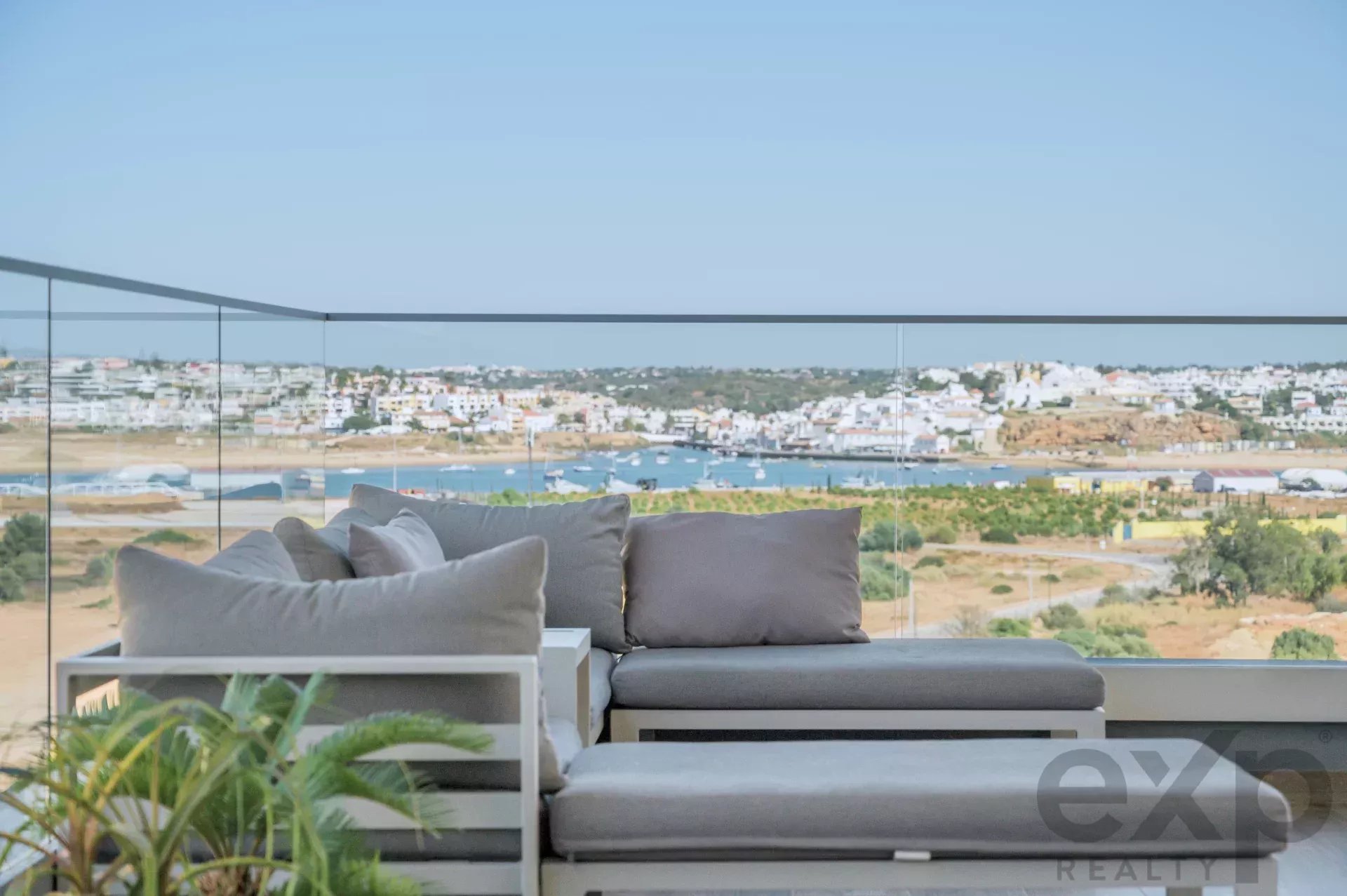 Glass-walled balcony with a gray-cushioned sectional sofa overlooking a coastal town and harbor.