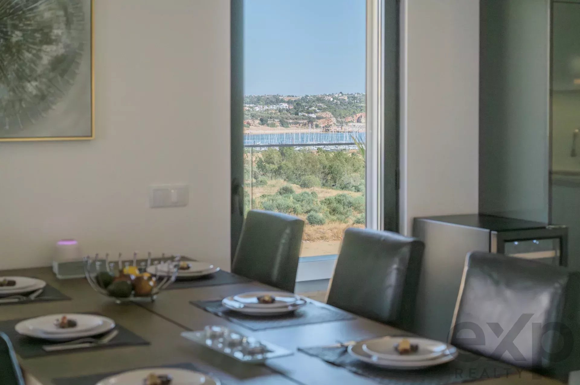 Dining room with a long table set for dinner, plates and glasses arranged, view of boats and hills through large windows outside.