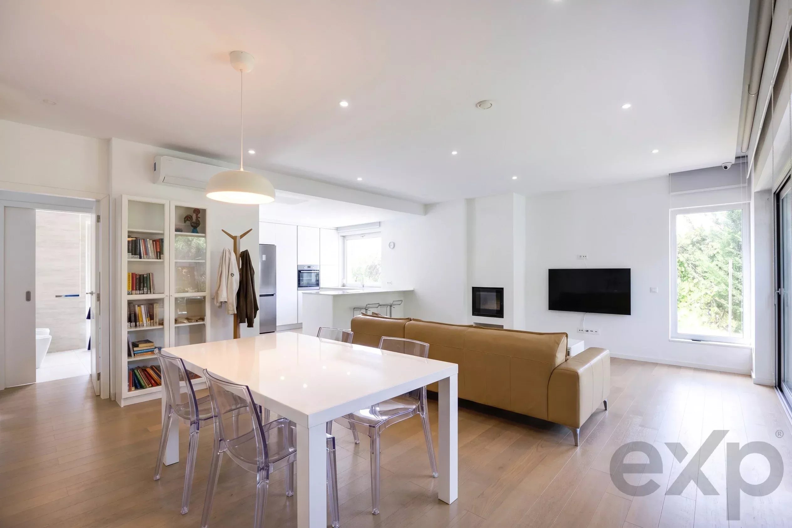Bright open-plan living area with white walls, a white dining table, transparent chairs, beige sofa, and a bookshelf-lined wall.