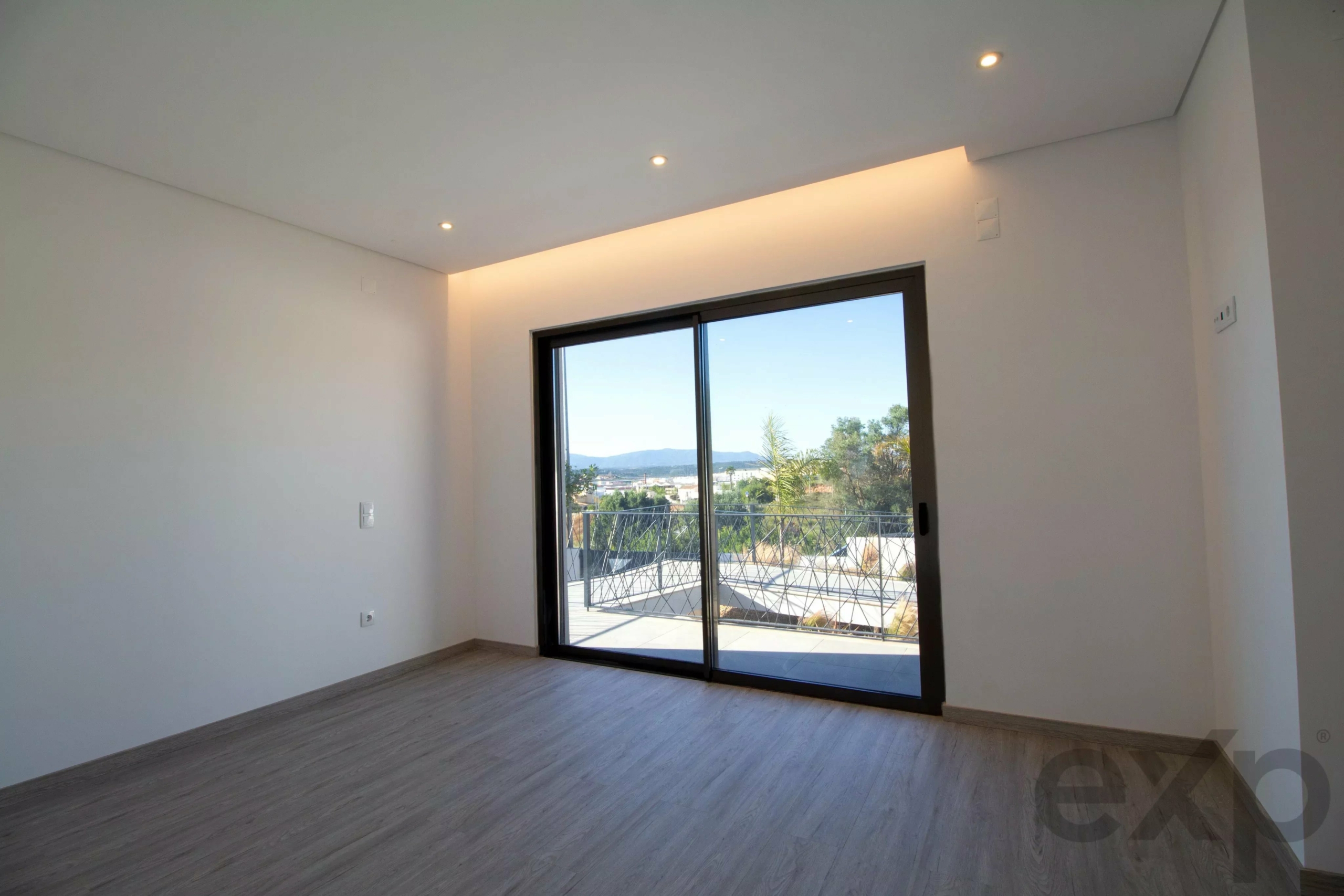 Empty modern room with a large sliding glass door opening to a balcony and hills outside behind a railing.