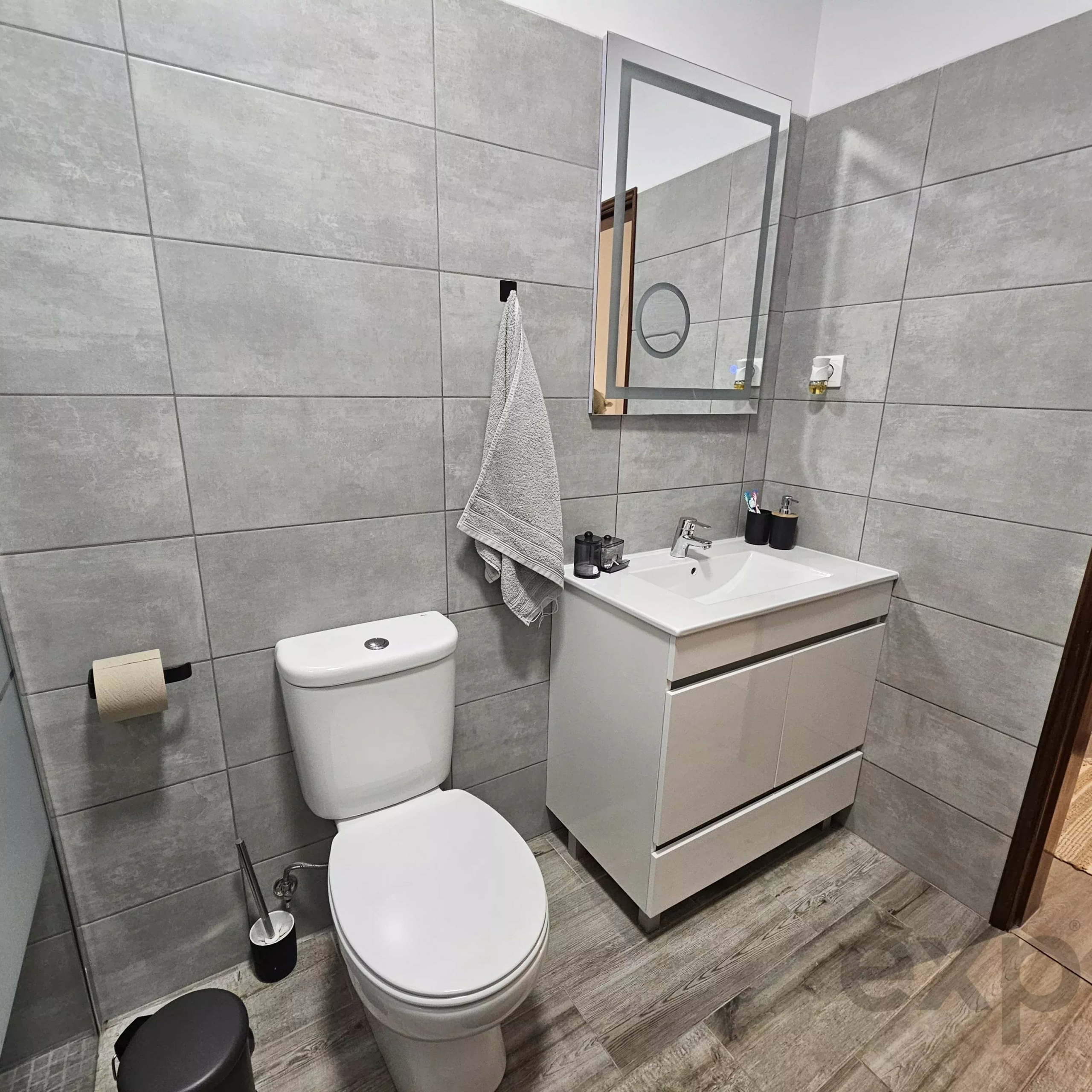 Modern bathroom with gray tile walls, a white toilet, and a white vanity with sink; towel hanging on wall.