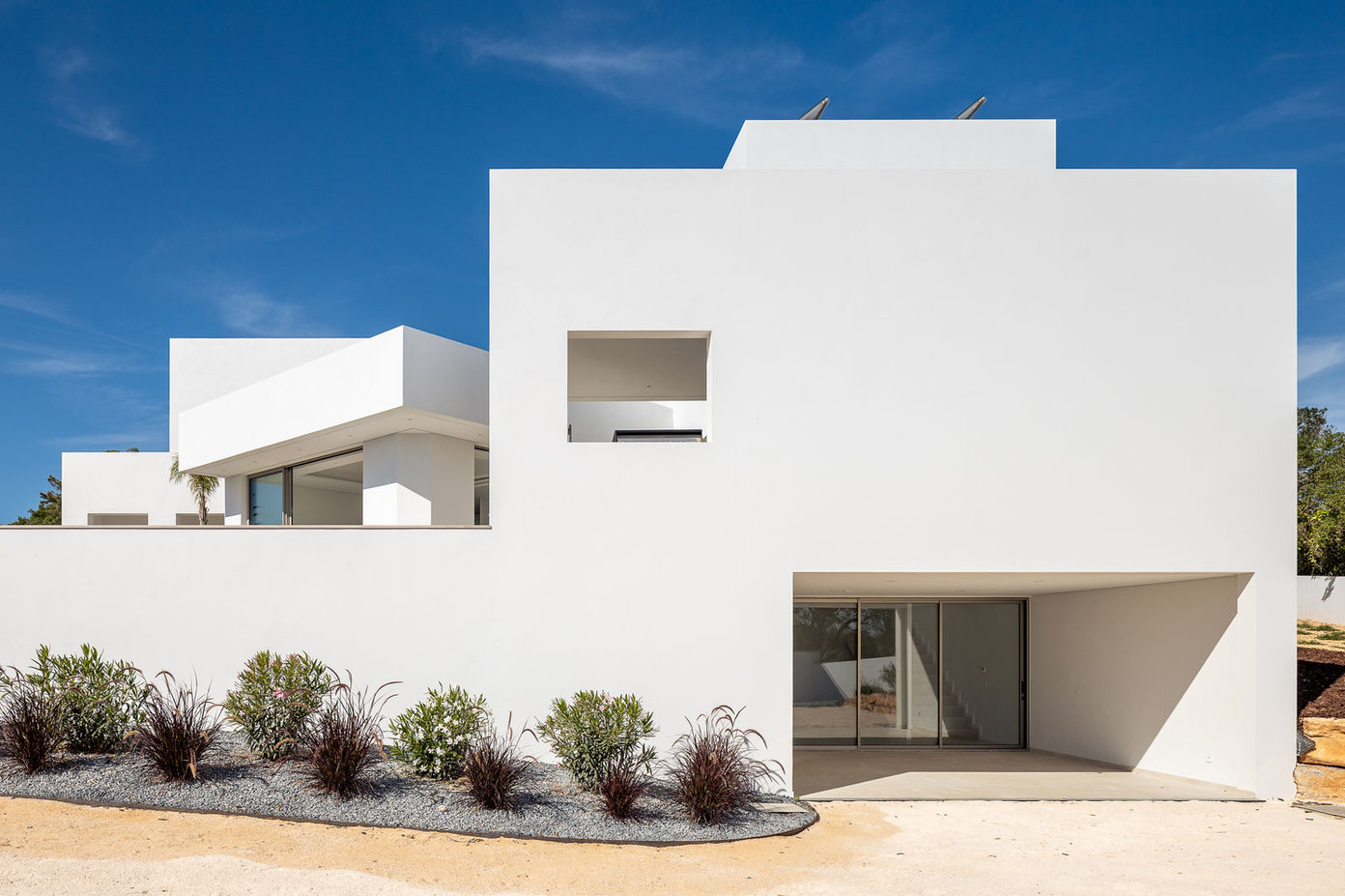 29bf52_183f258c98204c739e1ddbfe14664ad1-mv2 Modern white, boxy house with clean lines and large glass doors, set against a bright blue sky; desert landscaping in the foreground.