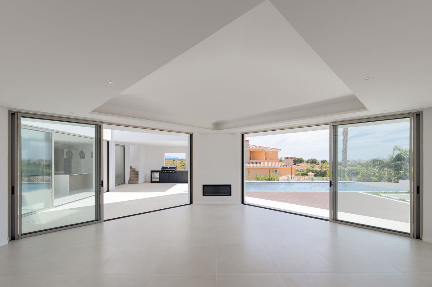 29bf52_28005f743fd54d99bdc36b140e505881-mv2 Bright open-concept living room with large sliding glass doors opening to a pool deck outdoor area and clear blue sky ahead, minimalist white interior in view