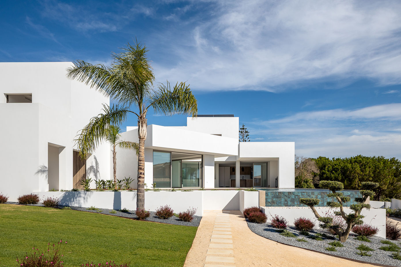 29bf52_33d38bd886cd4a0d986f022e3680ed4d-mv2 White modern house with geometric design, palm trees, and a rectangular pool set against a bright blue sky.