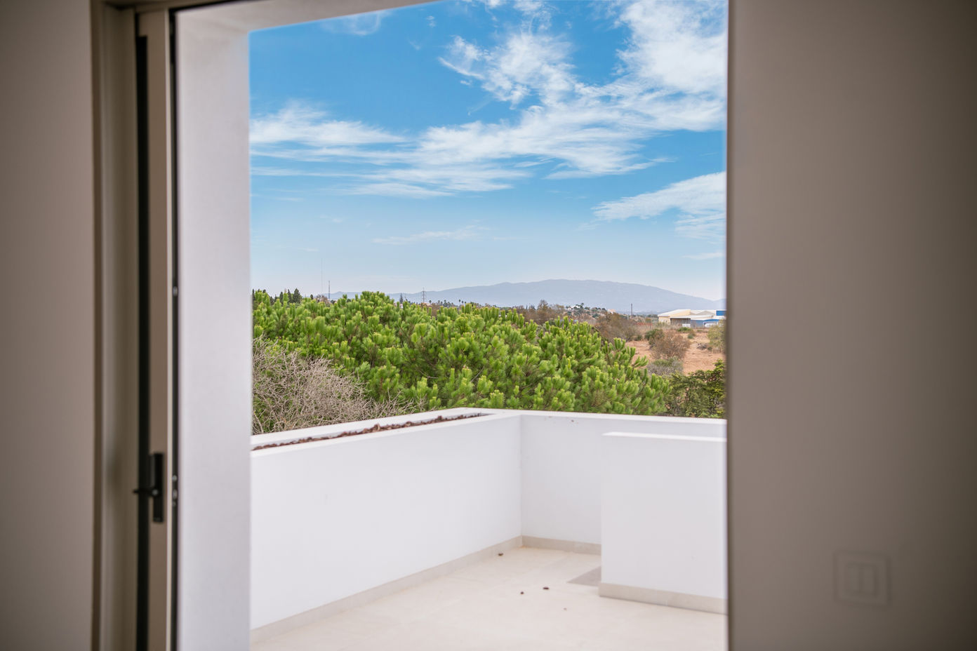 29bf52_34baeae5350c4dff9b06b09fe894c6c4-mv2 View from a balcony: white railing and floor, green trees beyond, with mountains and blue sky in the distance.