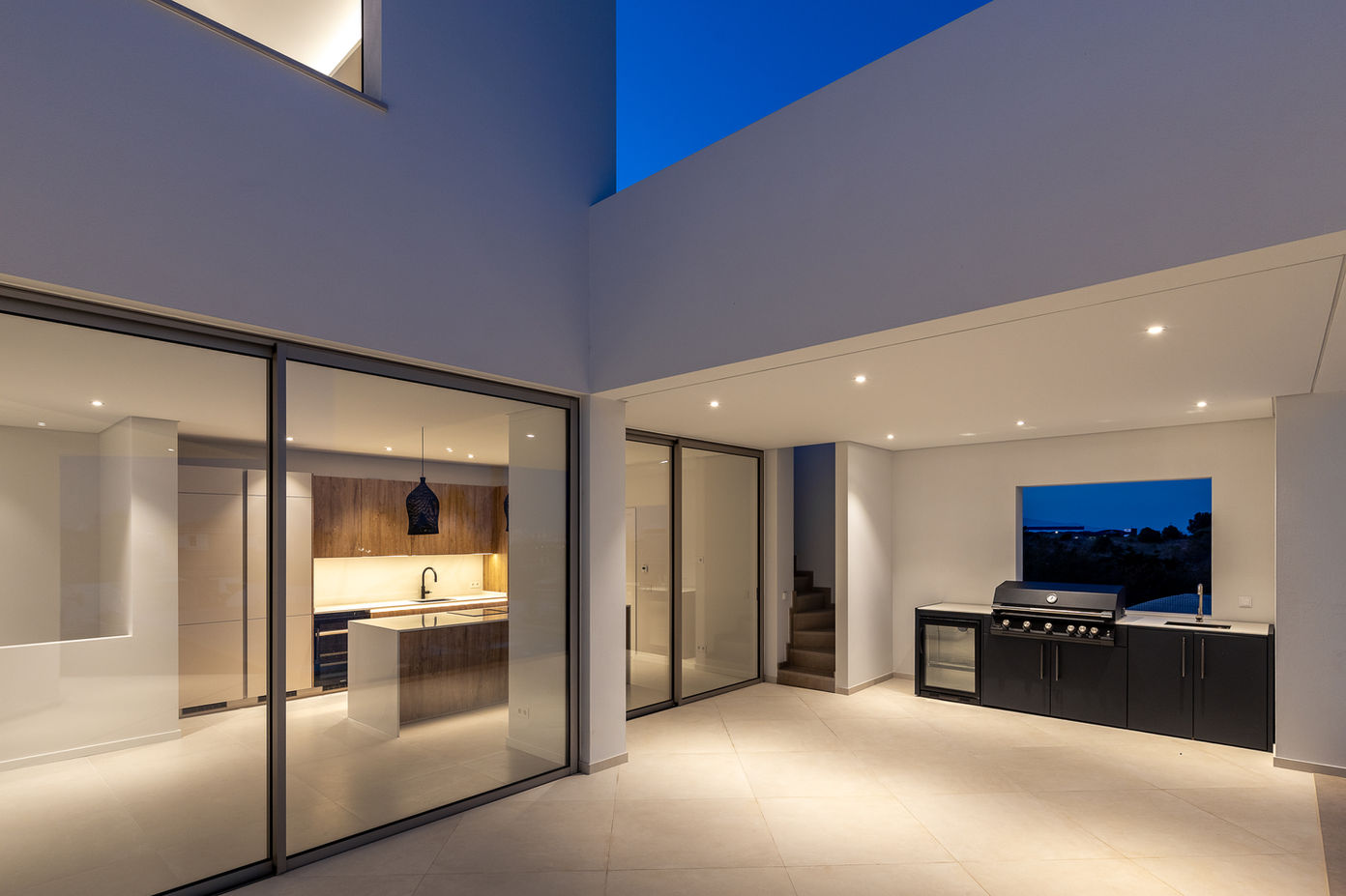 29bf52_3c018a864fa0445aa94701b3046c9adb-mv2 Modern outdoor patio at dusk with a built-in barbecue, kitchen island, and glass walls showing the lit indoor kitchen.