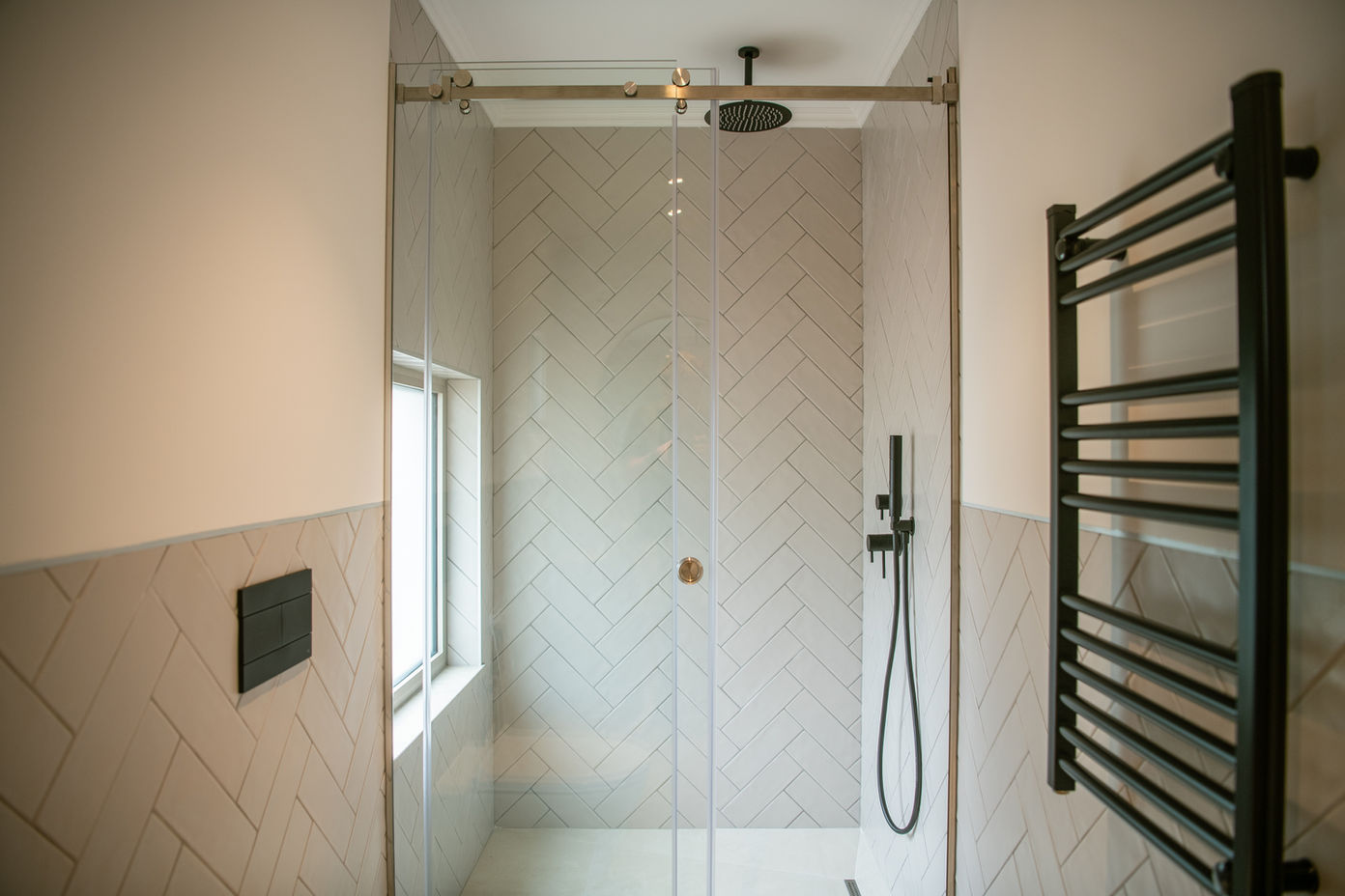 29bf52_3d7f1e53534145b1a10568a5755ea183-mv2 Modern glass-walled shower with herringbone tiled walls, rainfall showerhead, and black handheld shower on the right.
