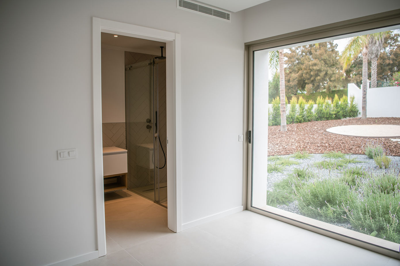 29bf52_4a62b77ec04a4416ad9f15d9fc9aec98-mv2 Bright, modern room with a large glass window overlooking a landscaped garden; doorway leads to a glass-enclosed bathroom with vanity visible inside.