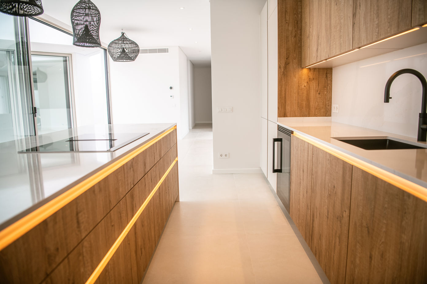 29bf52_5b13a7a5c4ac4b18abce657a1473efb4-mv2 Bright, modern kitchen with a long wooden island, white countertops, and black pendant lights overhead.