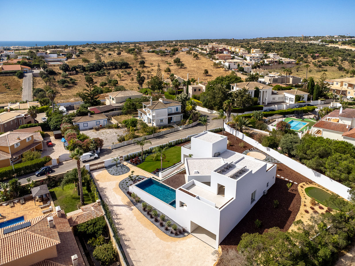 29bf52_64134b229772400ca9bf9b8fae45cd0d-mv2 Aerial view of a modern white villa with a rectangular pool, landscaped yard, and surrounding houses near a dry, coastal landscape.