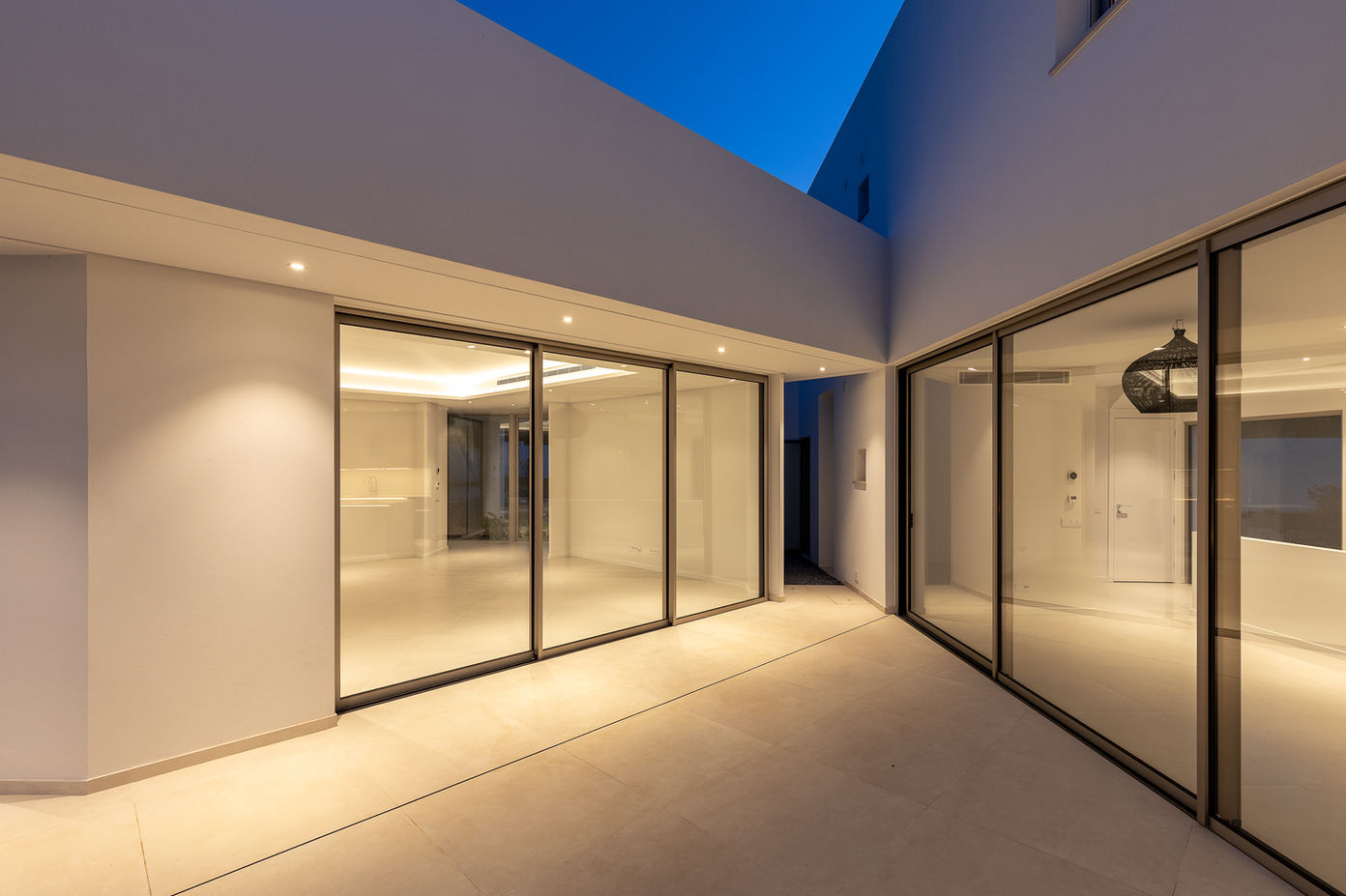 29bf52_7e3088ed78014588b123dd76212f8eec-mv2 Modern interior courtyard at dusk with expansive glass sliding doors and warm indoor lighting.