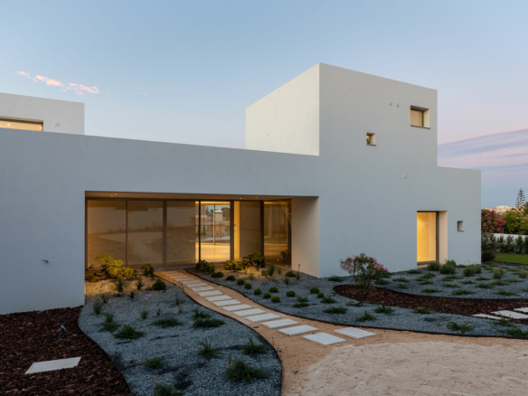 Modern white, minimalist house with large glass doors and a curved stepping-stone path through a desert-style garden, at dusk/often warm evening light.