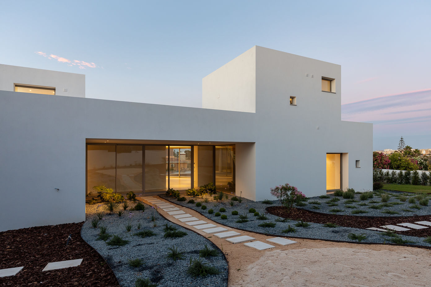 29bf52_830536e8da1048cbb237564f0a470e36-mv2 Modern white, minimalist house with large glass doors and a curved stepping-stone path through a desert-style garden, at dusk/often warm evening light.