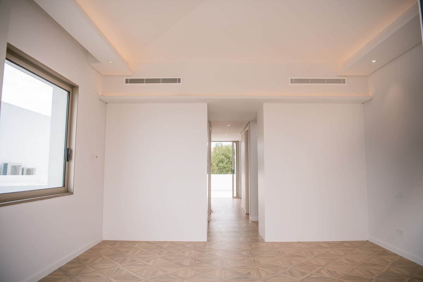 29bf52_95dbad6a8cb54ce9a454312afda09a3d-mv2 Bright, empty, modern hallway with white walls, a large left window, and a patterned wood floor leading to a glass door at the far end.