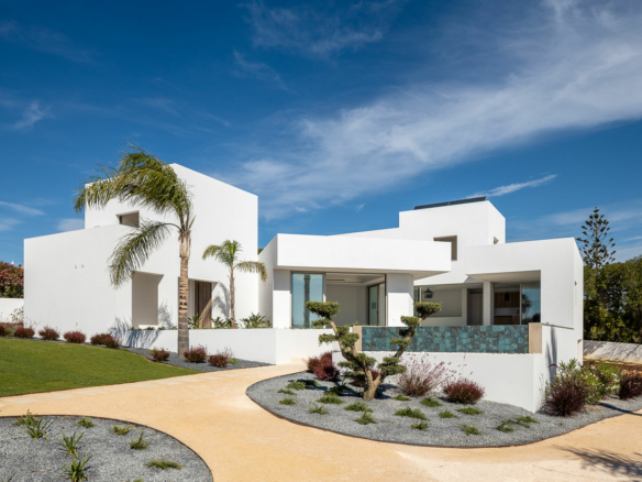 Modern white villa with angular blocks, palm trees, and a curved tan path in a sunny desert yard.