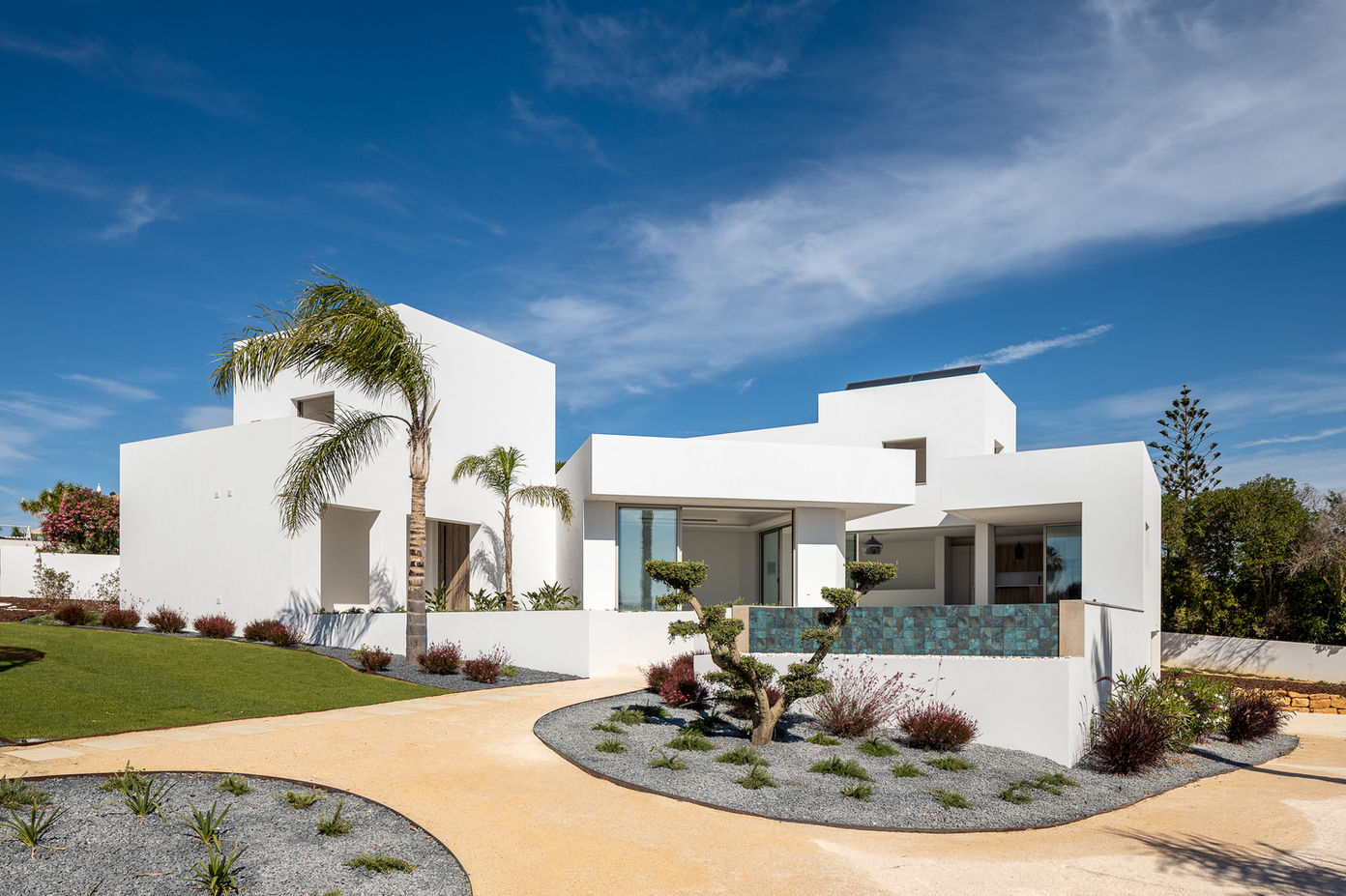 29bf52_99cb3ae3600e4a958cd8ba96a24ba89c-mv2 Modern white villa with angular blocks, palm trees, and a curved tan path in a sunny desert yard.