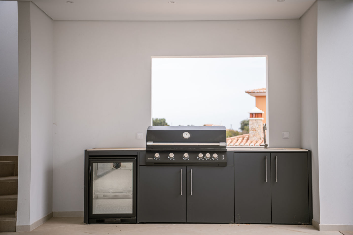29bf52_a24ca9f80e5d4f3282a2d148cb708f0e-mv2 Modern outdoor kitchen setup: built-in grill with a row of control knobs, small glass-door fridge to the left, and dark gray cabinets beneath a large window. A view of neighboring houses is outside the window.
